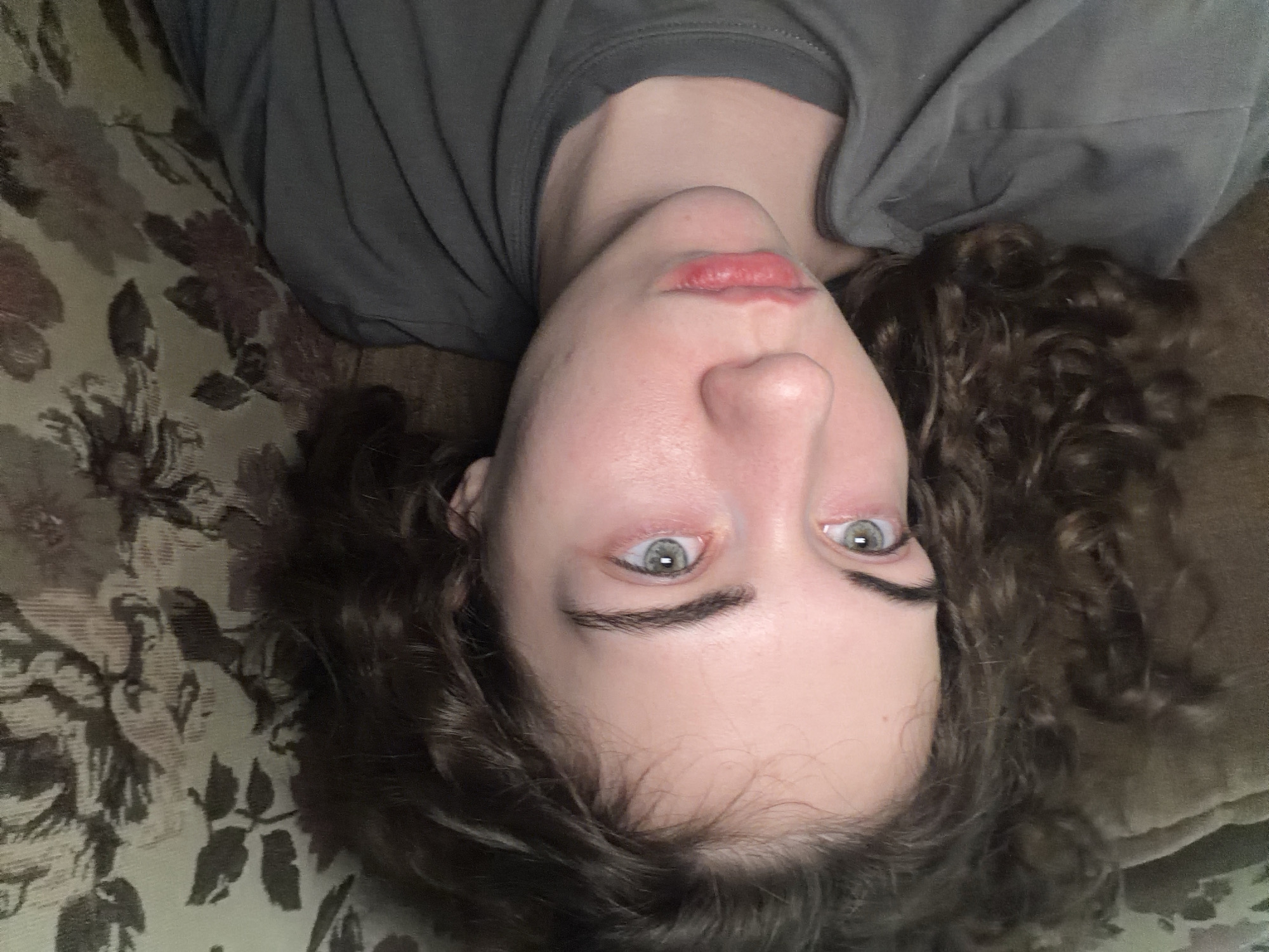 The image shows a close-up, overhead shot of a young woman lying on her back, with her head tilted slightly to her right. She has curly, dark brown hair that partially obscures her left side. Her skin is pale, with some redness around her eyes and cheeks. She has light green eyes and dark eyebrows. Her lips are slightly parted and appear to be colored with a reddish tint. She is wearing a dark gray t-shirt. She is lying on a surface that appears to be a couch or bed, covered with a patterned fabric. The pattern consists of dark, floral designs on a lighter background. The scene is indoors, with soft lighting. There is no indication of the city of Zavodoukovsk, Russia, or any specific location within it.