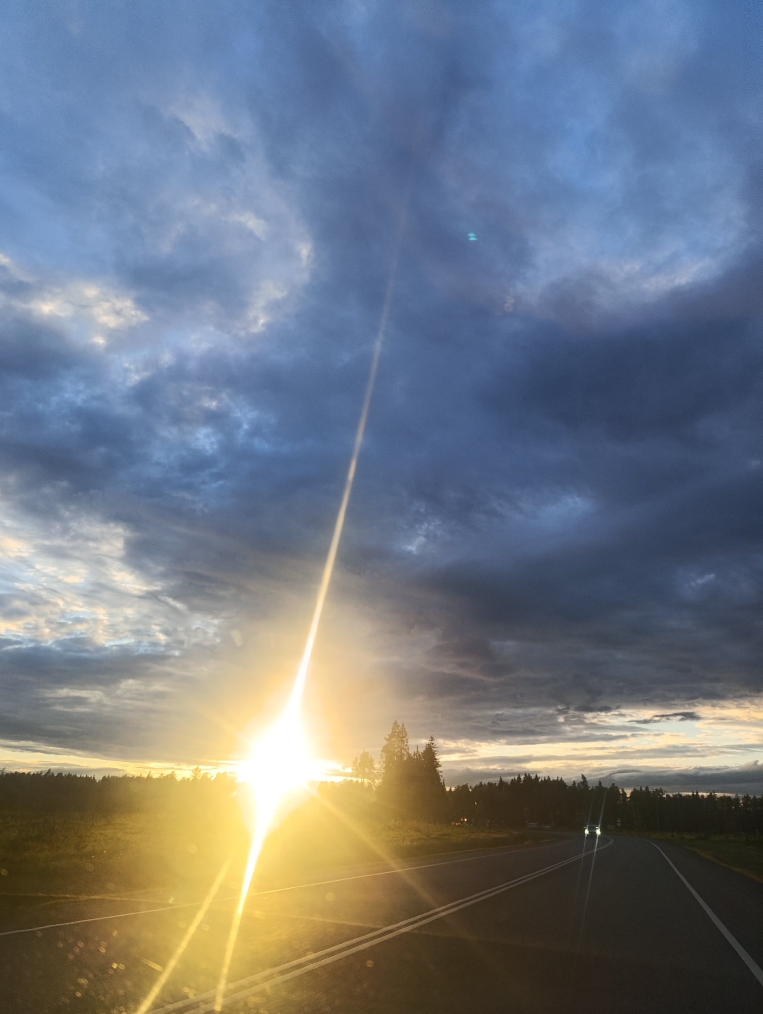 The image captures a road at dusk, bisected by double white lines, likely in a rural area. A car with headlights on is visible in the distance. The horizon is defined by a dark green tree line, which transitions into a bright, intense sun, creating a strong lens flare and casting a golden glow. The sky is a mix of dark, stormy clouds and patches of blue, suggesting a sunset or sunrise. The location is described as Unknown, Unknown.