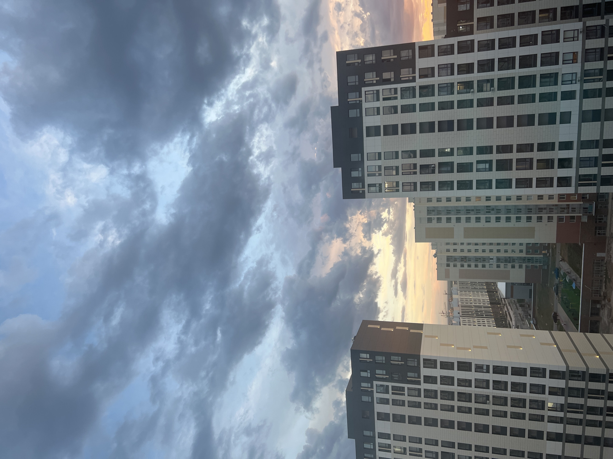 The image depicts a collection of modern high-rise apartment buildings under a dramatic, cloudy sky. The architecture features repetitive window patterns and a combination of light-colored facades with darker elements at the top. The sky displays a mix of dark gray and lighter, possibly sunset-tinged clouds, suggesting either dawn or dusk. In the foreground, two prominent buildings dominate the frame, with more structures stretching into the background, indicative of urban development. The ground level shows hints of infrastructure like roads and perhaps some green spaces or construction zones. The scene is set in Astana, Kazakhstan, as indicated by the context. There are no people clearly visible in the image, and the focus is on the architectural landscape and the atmospheric conditions of the sky.
