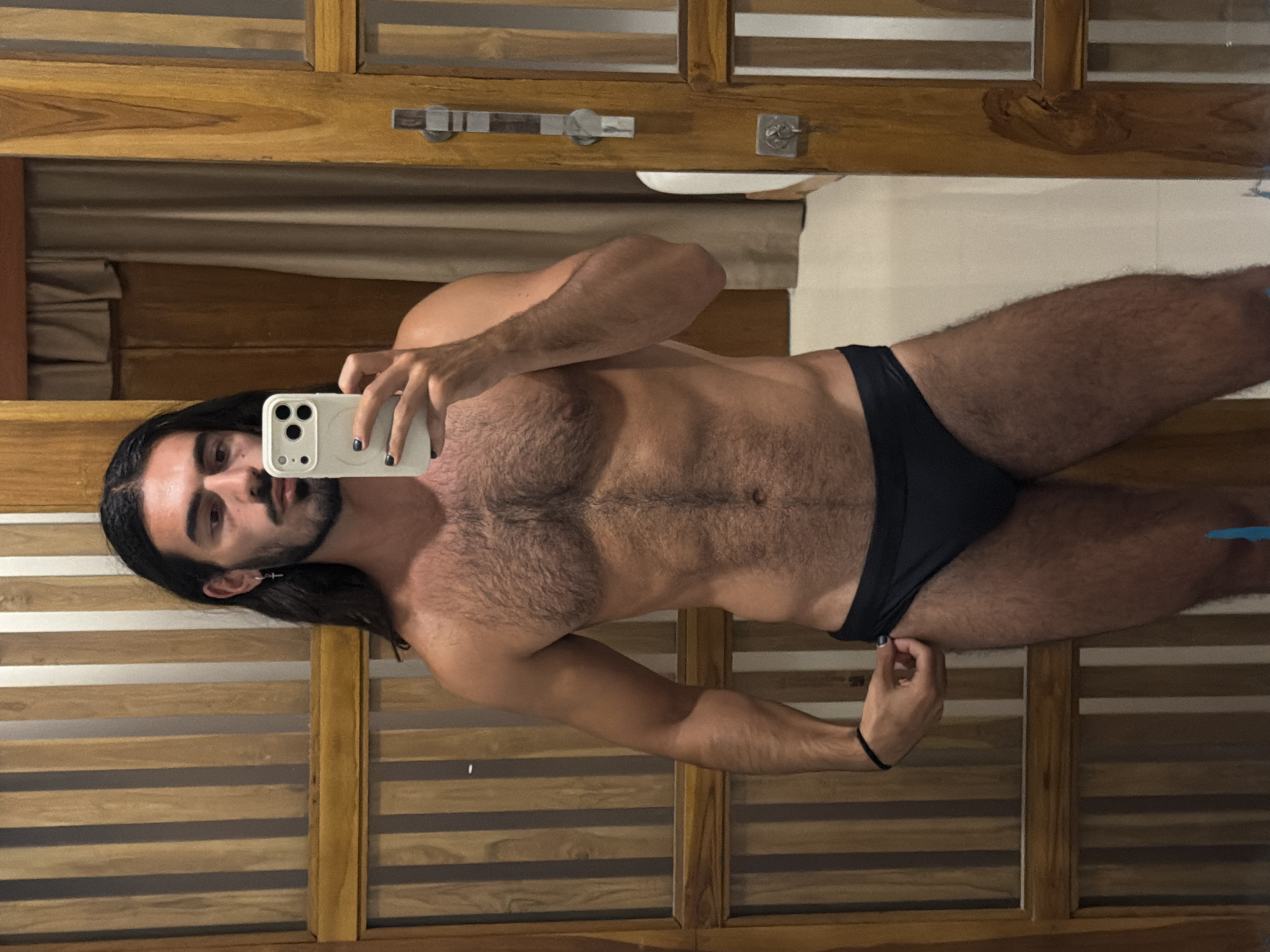 A person is taking a mirror selfie in a bathroom. The person has long dark hair and a beard, and is wearing black swim briefs. They are holding a white smartphone in their left hand. The background appears to be a wooden wall with horizontal slats and a beige curtain. A silver towel rack is visible on the wall. The lighting suggests it is daytime, possibly indoors. The overall impression is a casual, personal moment.
