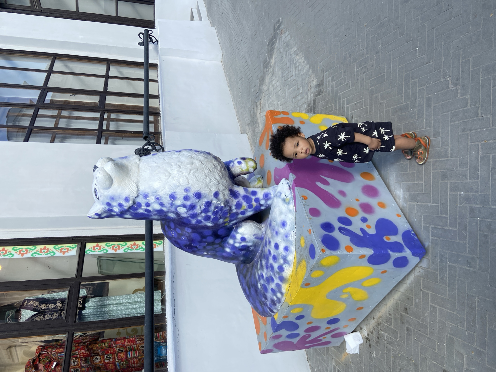 A young child lies on a colorful, cube-shaped object next to a stylized, purple-spotted owl sculpture on a gray-brick sidewalk in Kazan, Russia. The child wears a dark shirt with white palm tree patterns and brown sandals. A white building is in the background with windows displaying various fabrics. A black, decorative street lamp stands near the owl sculpture.