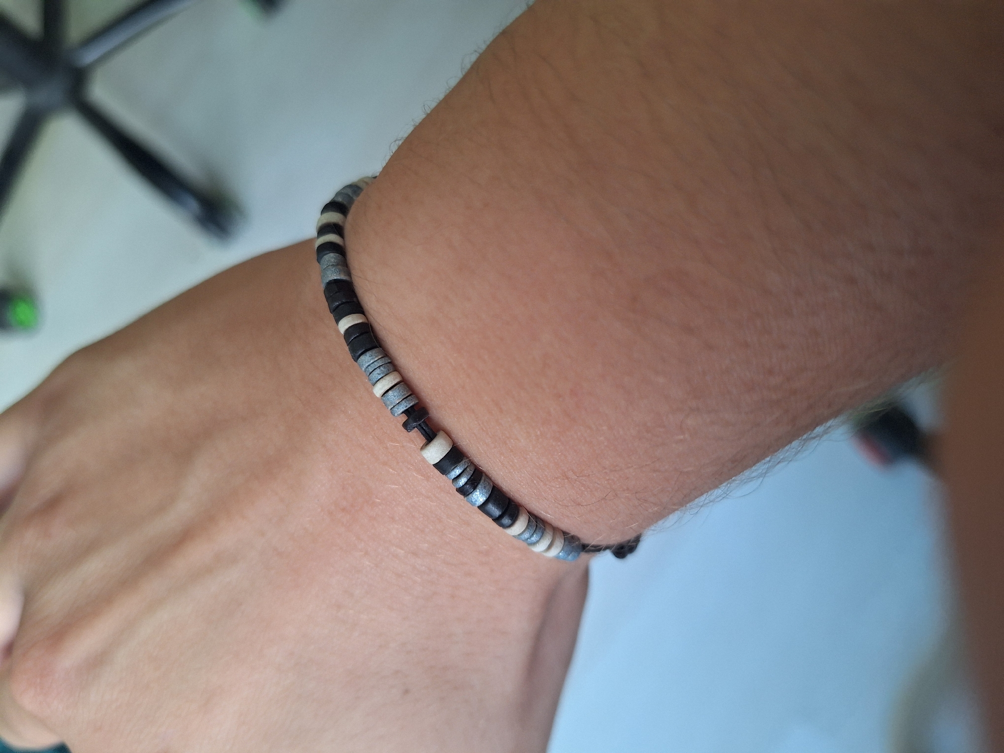 The image shows a close-up of a person's arm and wrist wearing a beaded bracelet. The bracelet is made of small, flat, disc-shaped beads in alternating colors of black, white, and gray. The background is blurred, but appears to be an indoor setting with a light-colored surface and the leg of a black office chair with green accents. The location is specified as the city of Varna, Bulgaria.