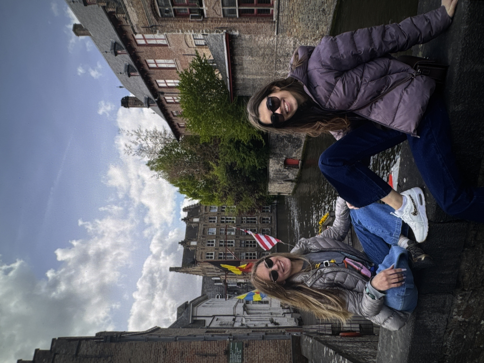 Two young women, both wearing sunglasses and puffer jackets, are sitting on a stone wall. The woman on the left has long blonde hair and is wearing a silver jacket over a gray t-shirt. The woman on the right has long brown hair and is wearing a purple jacket. They are both smiling. They are seated in front of a canal lined with brick buildings and flags. The architecture suggests a European city. This scene is not located in Kaunas, Lithuania.