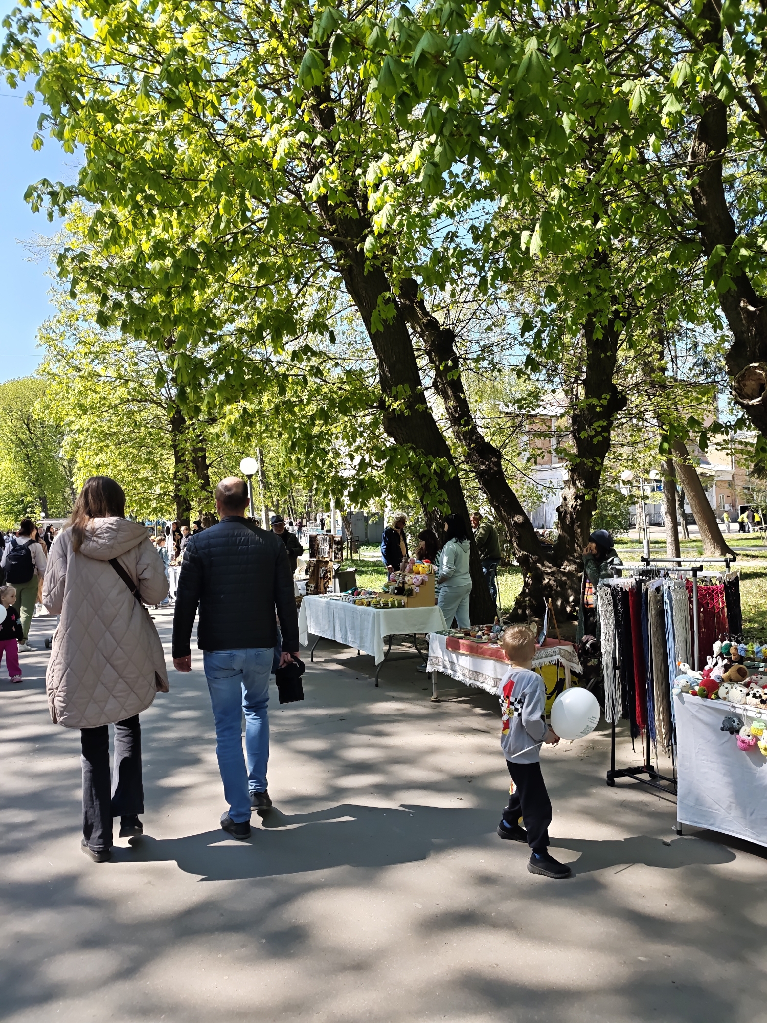 The image depicts an outdoor scene in Vinnytsia, Ukraine, likely within Central Park. The photograph is taken at eye level, capturing a wide walking path or street bordered by mature trees, several of which are in full green leaf.
A couple is walking away from the camera, the woman on the left wearing a beige coat and dark pants, and the man wearing a black jacket and blue jeans. Slightly ahead and to the right, a young boy is seen holding a white balloon.
Several stalls or tables displaying various goods are set up along the side of the path, suggesting an outdoor market or fair. The merchandise includes craft items, colorful knitted goods, and other handmade wares. A few people are browsing or interacting with the vendors at the stalls.
In the background, other pedestrians can be seen walking in both directions, and buildings are visible in the distance. The sky is a clear, light blue, and the bright sunlight casts shadows on the ground. The overall atmosphere appears lively and pleasant.
