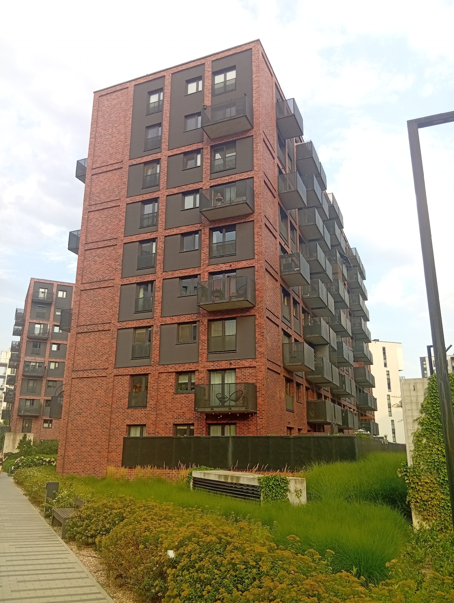 A low-angle, full shot captures the exterior of a modern apartment building in Wrocław, Poland. The building is constructed primarily of red brick, with dark grey rectangular panels interspersed between floors, and features numerous dark grey balconies with glass railings. The building's facade is dominated by a grid of windows and balconies. To the left, a second, similar building is partially visible, also featuring balconies and brickwork. The foreground consists of a paved walkway with a grey and white patterned design, bordered by lush green and yellow-green foliage. A wooden bench is situated on the left side of the walkway, amidst the greenery. In the background, other modern buildings, including a tall white structure, are visible. The sky is overcast with scattered clouds. A tall, dark, rectangular lamppost is present on the right side of the image.