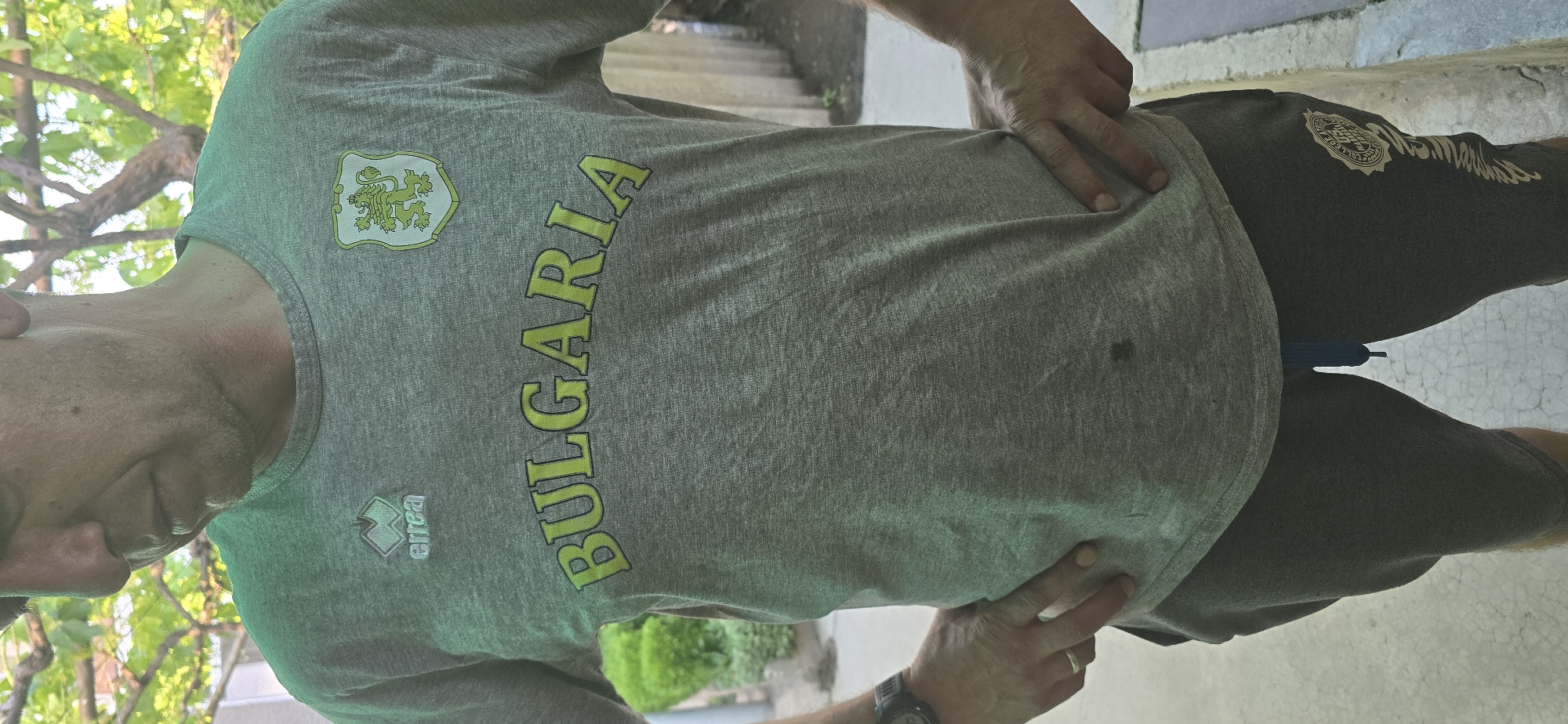 A person, likely male, stands outdoors in a casual, relaxed pose. The individual is wearing a grey marl t-shirt and dark grey shorts. The setting features green foliage and tree branches in the background, indicating a daytime scene. The ground is a light-colored, cracked paved or concrete surface.
The grey t-shirt prominently displays national and brand identifiers. On the upper left chest, a crest-like emblem depicting a stylized lion, characteristic of the Coat of Arms of Bulgaria, is visible. Below this, the word "BULGARIA" is emblazoned in large, bright green capital letters with a darker outline across the chest. The "errea" sportswear logo is also present below the crest on the left side.
The person's hands are placed on their hips, and a watch is worn on the left wrist. A small blue object, possibly a pen or marker, is tucked into the right side of the shorts' waistband. The dark grey shorts feature a circular logo on the right thigh, which appears to include partial text like "COLLEGE" and "MARSHALL". The person's face is partially visible in profile, showing some stubble, but no distinct emotion is discernible. The overall scene suggests a casual moment in Govedare, Bulgaria.