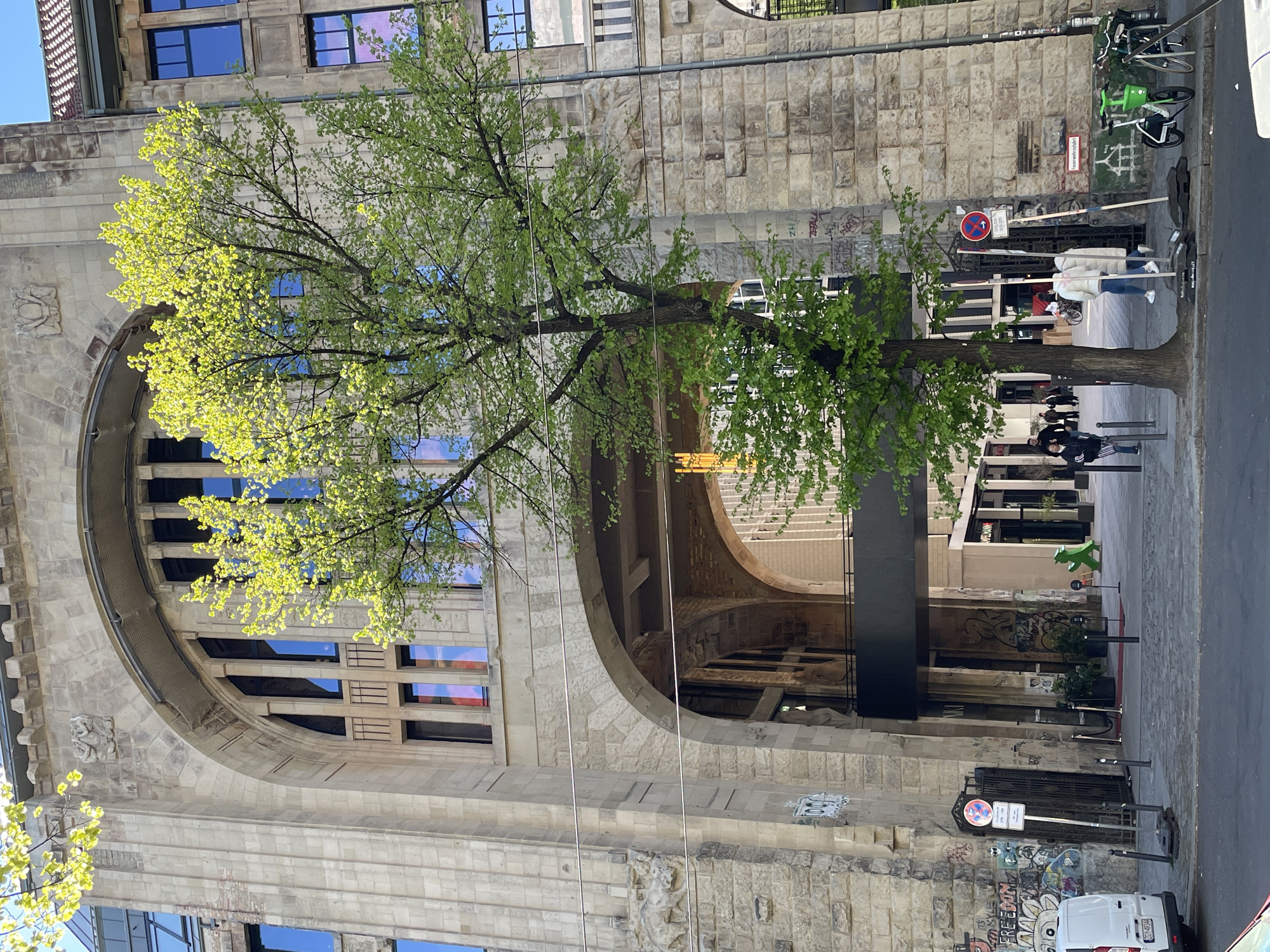 This image depicts a sunny urban street scene, likely in Berlin, during springtime. The primary subject is a grand, multi-story stone building with a traditional architectural style, featuring large, cut stone blocks and ornate carvings, including a relief sculpture. A prominent, deep archway with internal structure is visible within the building's facade.
In the foreground, a deciduous tree stands with vibrant, newly-sprouted light green leaves, indicating the season is spring. The tree's canopy extends across much of the left and central parts of the image, partially obscuring the building's upper windows, which reflect the clear blue sky.
On the right side, a paved sidewalk or street recedes into the distance. Several people are visible on the sidewalk, walking or standing, suggesting everyday activity. Various objects are present, including several bicycles parked against the building's stone wall, some with green frames. The lower portions of the stone walls and surfaces on the right show scattered graffiti.
Street furniture includes multiple dark bollards and several signs. A prominent red and blue "No Parking" sign is clearly visible, beneath which is a white rectangular sign displaying the German word "Einbahnstraße" (meaning "One-way street"). Further along the sidewalk, a distinctive green, abstract sculpture, possibly resembling a dinosaur, stands near a commercial entrance. A white van is partially visible in the bottom right corner. The strong sunlight and clear reflections in the windows indicate a bright, sunny day.