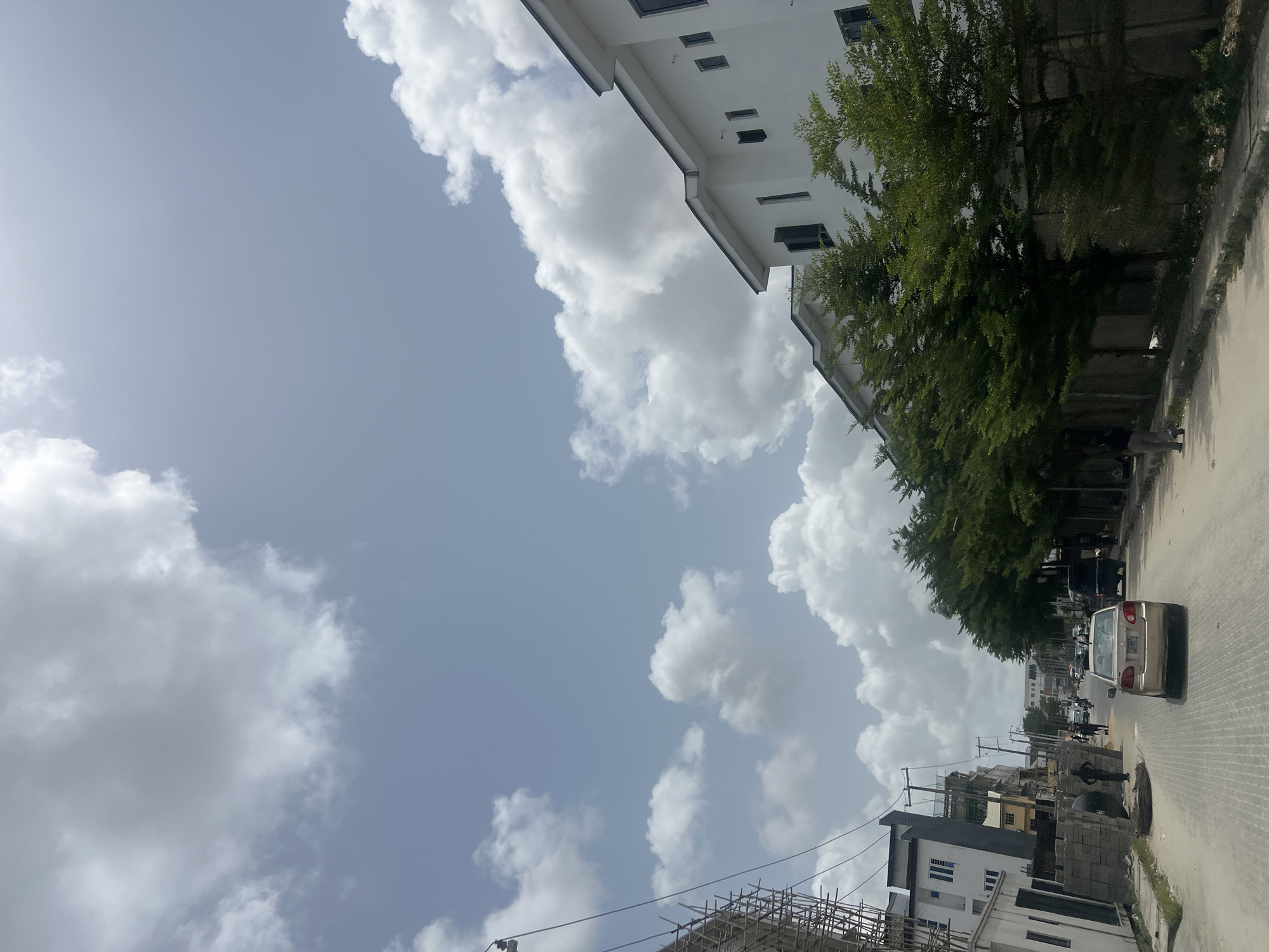 This media file captures a vibrant street scene in Igboefon, Nigeria, under a partly cloudy sky during what appears to be daytime. The image is taken from a low angle, looking upwards and along the street, emphasizing the expansive sky and the structures lining the road.
The upper portion of the frame is dominated by a pale blue sky dotted with large, fluffy white cumulus clouds. A soft sun glare is visible in the upper left corner, suggesting bright daylight.
Along the street, modern white buildings with multiple stories and dark, rectangular windows are prominent. These structures suggest a contemporary urban or suburban development. A dense line of green trees runs along the sidewalk in front of some of these buildings, providing shade and a natural canopy.
On the street itself, a light-colored sedan, possibly gold or beige, is visible on the right side of the road. Other vehicles are further down the street, though less distinct. A few people are present on the street; one figure can be seen near a construction site in the foreground, and others are further in the distance.
The foreground also features signs of ongoing construction, including scaffolding made of wooden poles and stacks of concrete blocks, indicating development in the area. Street poles with overhead wires are also visible, characteristic of urban infrastructure. The road appears to be paved with stones or bricks.
Overall, the image portrays an active, developing street in Igboefon, characterized by modern architecture, green foliage, and daily activity under a bright, cloud-filled sky. No visible text is discernible in the image.