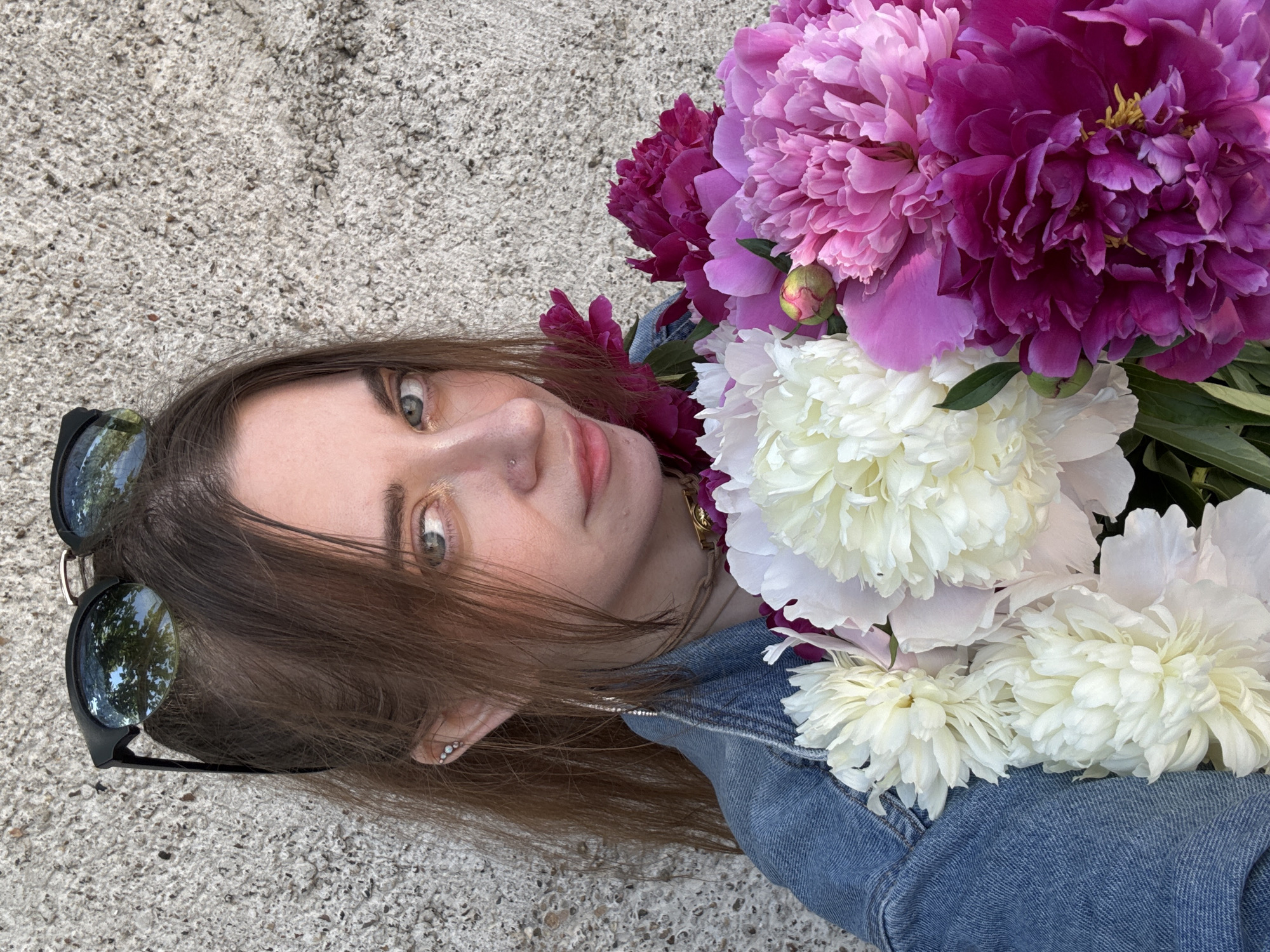 A young woman with brown hair, wearing sunglasses on her head, is holding a bouquet of peonies. She is wearing a denim jacket. The background is a rough, textured concrete wall. The image is a close-up shot, with the woman's face and the flowers filling most of the frame.