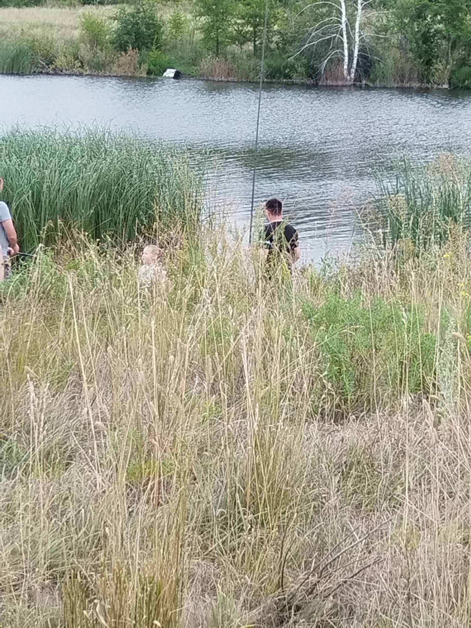 The image shows a body of water with reeds along the shore. A person in a black shirt stands facing the water, with a fishing rod. Another person, a child, is partially visible in the reeds to the left of the person fishing. A partially submerged object, possibly a bin, is visible in the water on the far side. The environment appears to be a natural area, with trees and vegetation in the background. It is not possible to determine the specific location within the city of Parfenovka, Russia based solely on this image.