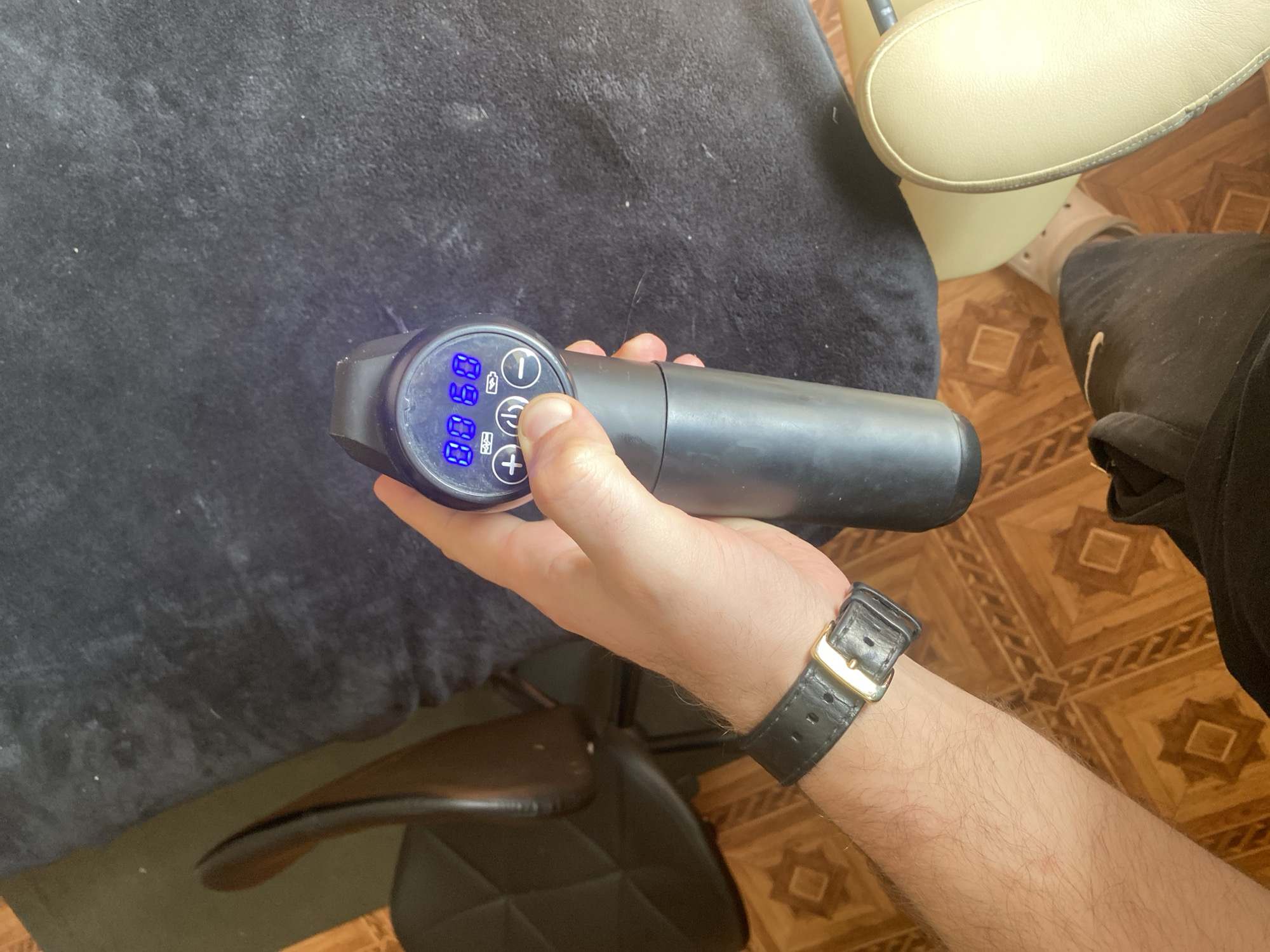 The image shows a close-up of a person's hand holding a black massage gun. The massage gun has a digital display with blue illuminated numbers that read "00 00". There are also visible buttons on the massage gun, including a power button with a circle and a line, and a plus button. The person is wearing a black leather watch with a gold buckle on their left wrist.
The background consists of a black textured surface, likely a blanket or fabric, and hints of a chair and wooden flooring. The lighting suggests it is daytime, but there are no specific indicators of the time of day or weather conditions. The overall scene appears to be indoors, possibly in a personal setting. The focus is on the massage gun and the hand holding it, implying the activity of using or demonstrating the device. There are no discernible emotions conveyed by the image. No other text is visible besides the numbers on the massage gun display.