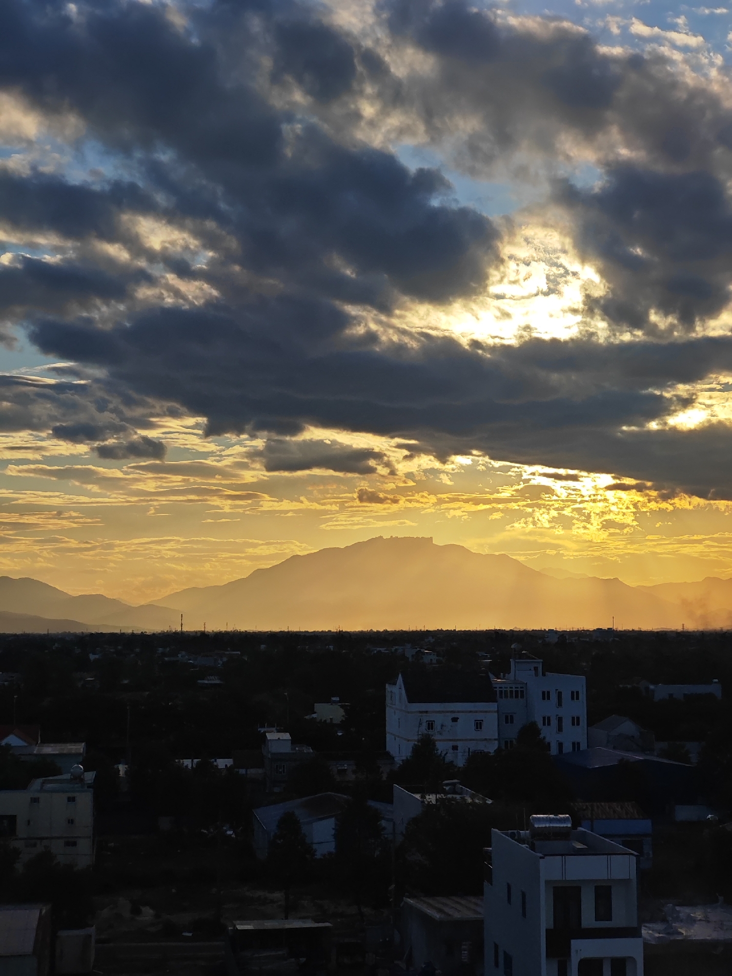 This image captures a panoramic view of a cityscape at dusk, likely in Đà Nẵng, Vietnam. The sky is dramatic, filled with a mix of dark, heavy clouds and bright patches where the setting sun is breaking through, casting a warm, golden glow. The sun's rays create a luminous haze that softens the distant mountain range silhouetted against the horizon.
Below the sky, the city unfolds in a dense arrangement of buildings and trees. The structures vary in size and style, with some appearing modern and multi-story, while others seem smaller and more traditional. The overall lighting suggests that the sun is low in the sky, casting long shadows and making the foreground buildings appear dark and silhouetted. The vegetation is a deep green, interspersed with the darker shapes of the buildings. The presence of what appear to be telecommunication towers indicates a developed urban environment. The atmosphere appears somewhat hazy, possibly due to pollution or the atmospheric conditions of sunset in a tropical region.