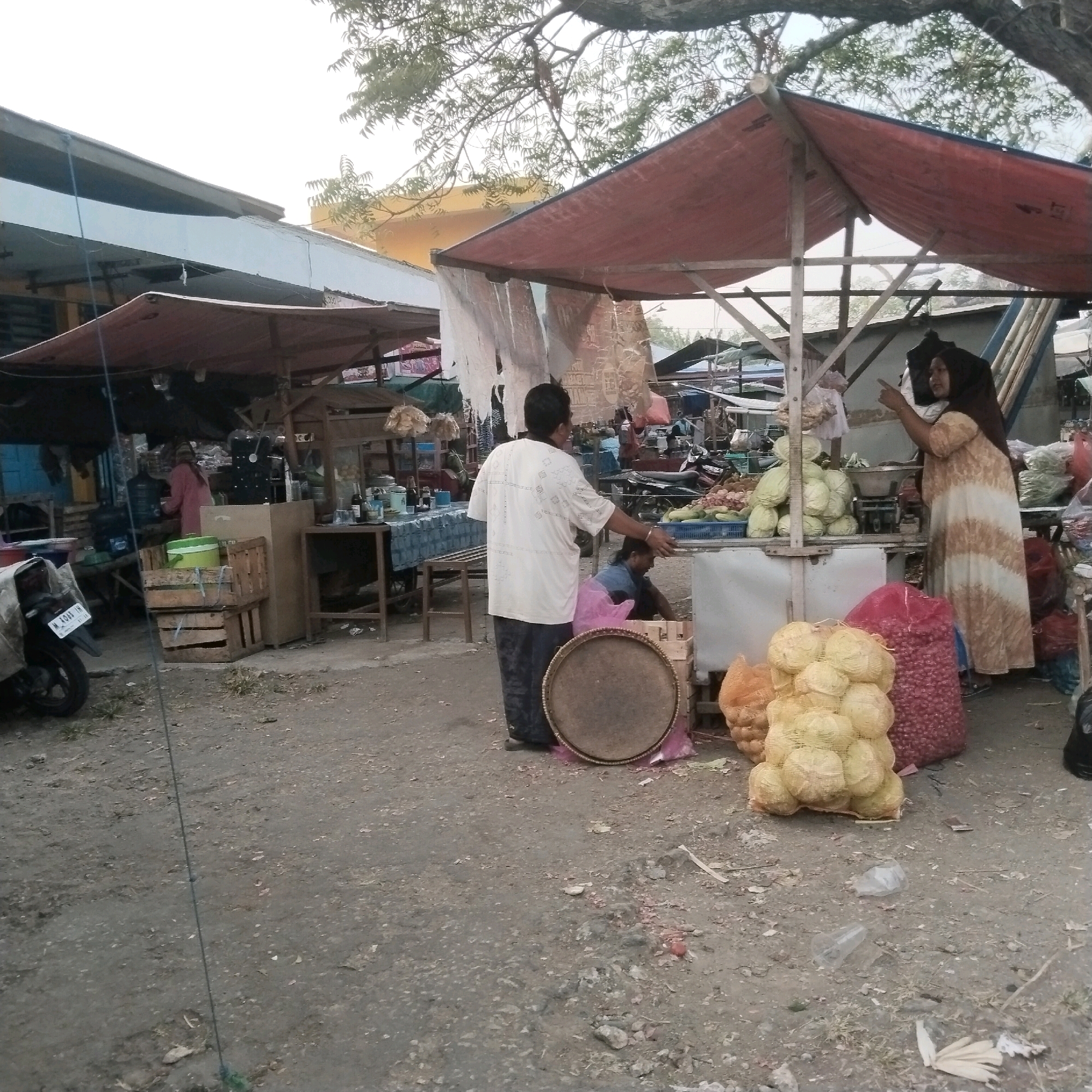 The image depicts an outdoor market scene in Kesek, Indonesia, characterized by a dusty, uneven ground and several temporary stalls. In the foreground, a motorcycle is parked and covered with a light-colored tarp. To the left, a stall is partially visible with wooden crates and a green bucket. In the center, a man wearing a white t-shirt with a patterned design and dark pants stands facing away from the camera, appearing to be interacting with someone at a stall. In front of him, a round, woven mat lies on the ground.
To the right, a woman in a patterned dress and a headscarf stands behind a stall, gesturing with her right hand. The stall displays various produce, including piles of cabbage and onions in mesh bags, and a blue crate of vegetables. Behind the woman, there is a scale and other market items. The background shows more stalls, trees with foliage overhead, and various structures, suggesting a busy marketplace. The overall atmosphere is that of a typical open-air market, likely in a rural or semi-urban area of Indonesia, given the location context. The lighting suggests it is daytime, possibly late morning or afternoon.