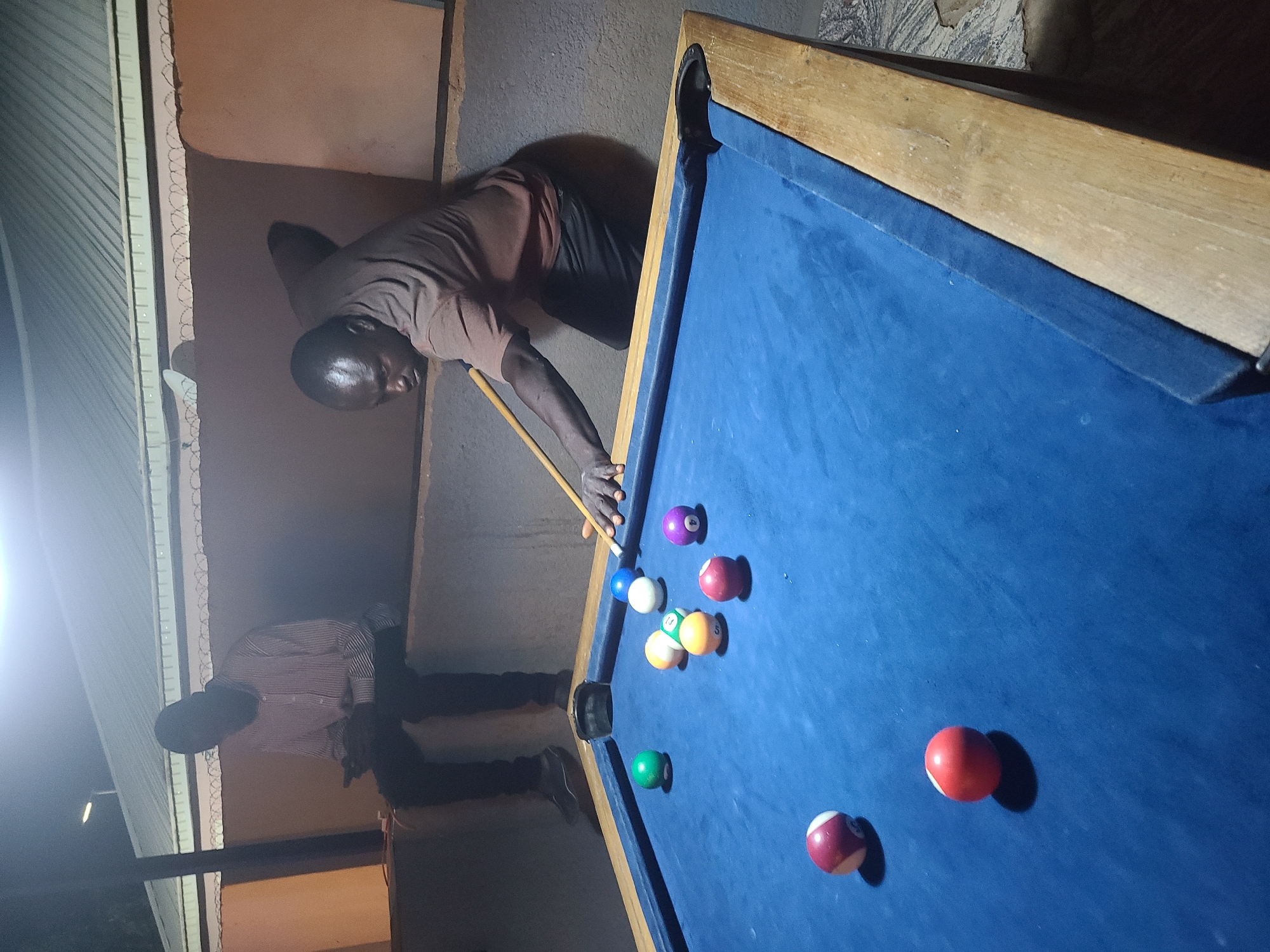 Two men are engaged in a game of pool (billiards) in what appears to be an indoor or semi-covered outdoor setting at night, as suggested by the artificial lighting and prominent shadows. The primary subject is a dark-skinned man in a brown t-shirt and dark shorts, leaning over a blue-felted pool table, holding a cue stick, and preparing to make a shot. His intense focus is evident.
To his left, another dark-skinned man wearing a striped shirt and dark trousers stands observing the game. The pool table, with its wooden rails and blue playing surface, is central to the image, covered with several colorful pool balls including solids (red, green, purple, yellow, blue, white cue ball) and striped balls. Strong shadows are cast by the players and the balls on the felt.
The background features rough-textured, earthy-toned walls, possibly concrete or plaster. On the far left, there's a corrugated metal structure, possibly a wall or roofing, and a section of barbed wire running along a low partition or wall, suggesting a practical, possibly informal, environment. The overall atmosphere is casual and focused on the game. No visible text is present. The setting appears to be a local establishment or gathering place in Gura Top, Nigeria.