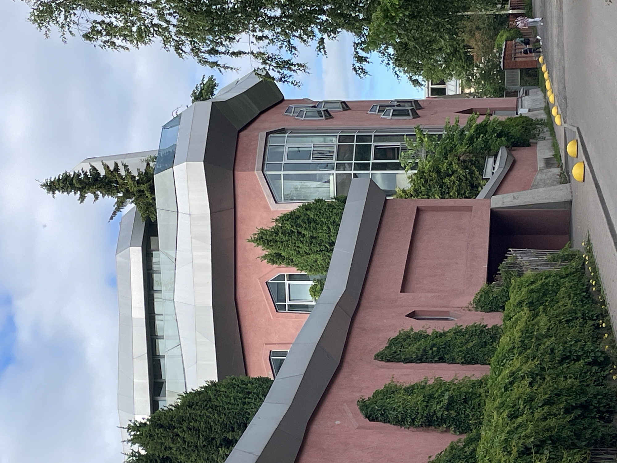The image depicts a uniquely designed building in Saint Petersburg, Russia, characterized by its pink facade and angular, multi-level roof structure. Several evergreen trees are strategically placed around the building, some appearing to grow from its upper levels. The building features numerous windows, including a prominent, multi-pane bay window on the right side, and several smaller, angled windows on the same facade.
The foreground shows a paved walkway or street lined with yellow, dome-shaped bollards. To the left, a dense green hedge and climbing ivy cover a lower section of the building, partially obscuring an entranceway. In the background, beyond the main building, other structures and more trees are visible. Two individuals, appearing to be a woman and child, are walking away from the camera on the right side of the paved area. The sky is partly cloudy, with a mix of blue sky and white clouds.