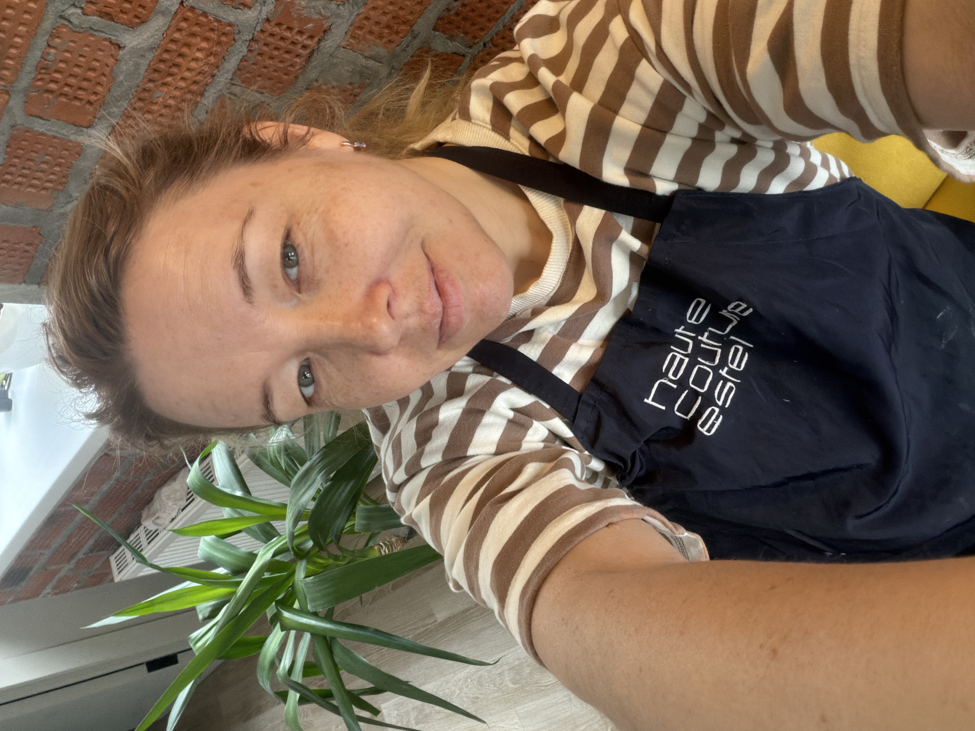 A woman with fair skin and freckles takes a selfie indoors. She wears a brown and white striped shirt and a dark apron with white lettering. She gazes directly at the camera with a slight smile. A green plant occupies the lower left foreground. In the background, a brick wall is visible at the top, and a window is suggested by the white frame at the left edge. The location in the city of Pervomaysky, Russia is not discernible from the image.