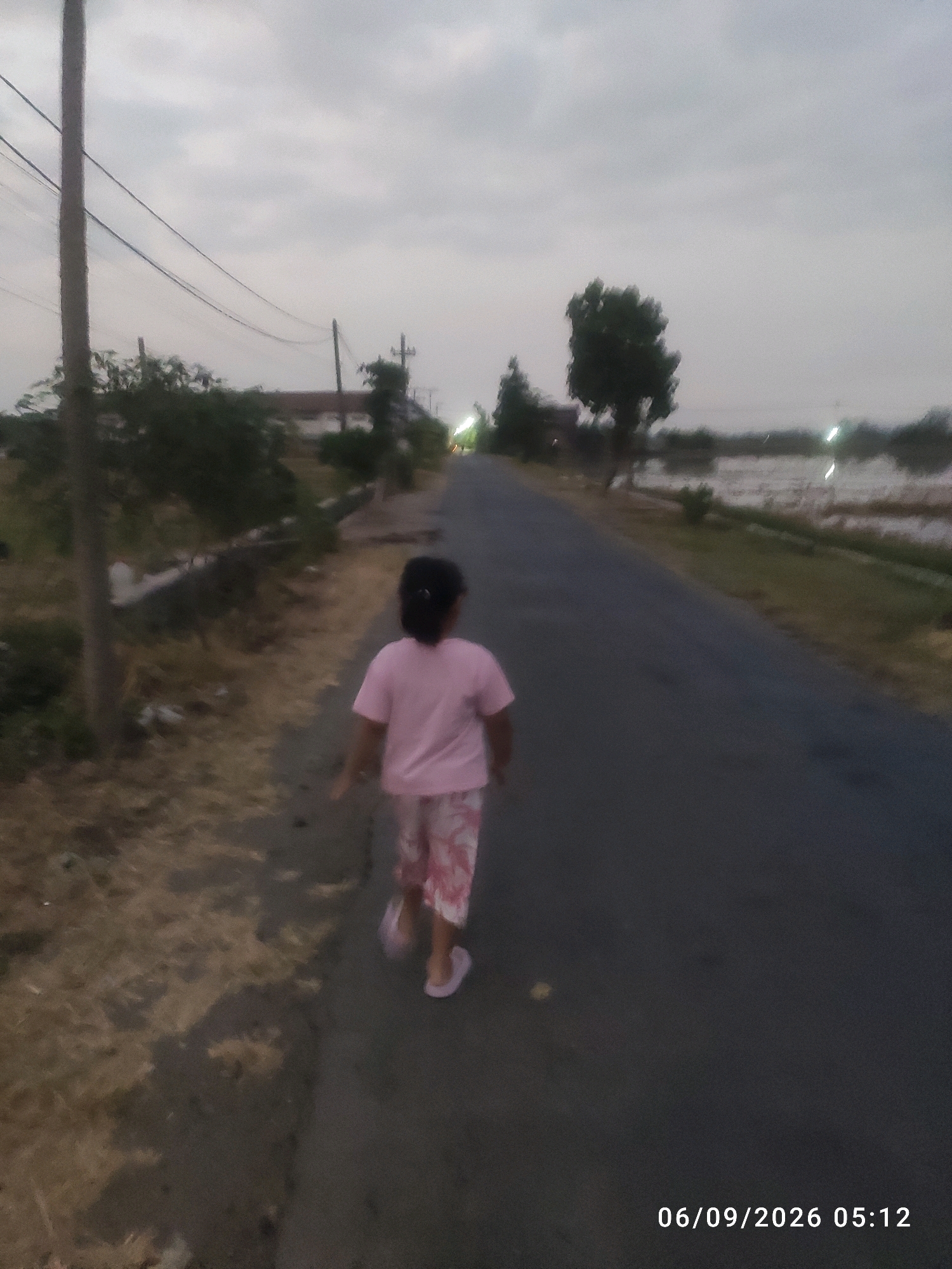 A young girl in a pink t-shirt and patterned pants walks away from the camera on an asphalt road. She is wearing pink flip-flops. The road is bordered by dry, overgrown grass on the left and a field of water on the right. In the background, there are trees, a building, and utility poles with wires. The sky is overcast. A timestamp in the lower right corner reads "06/09/2026 05:12". The scene appears to be in a rural or suburban area, possibly in Bongkot, Indonesia.