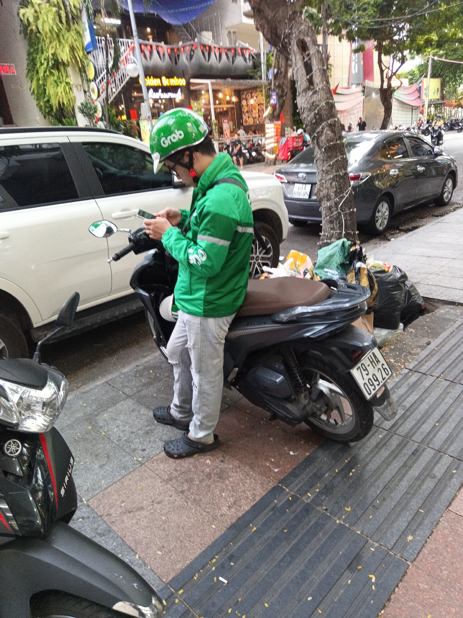 A man wearing a green "Grab" helmet and jacket is standing next to a black scooter on a city sidewalk in Nha Trang Ward, Vietnam. He is looking down at a smartphone. The scooter has a license plate that reads "79-HA 099.26". Next to the scooter, on the sidewalk, there are black garbage bags. A white car is parked behind the scooter, and a gray car is parked further down the street behind a tree with lights wrapped around its trunk. A portion of another scooter is visible in the lower left corner of the frame. The "Golden Bamboo Restaurant" is visible in the background.