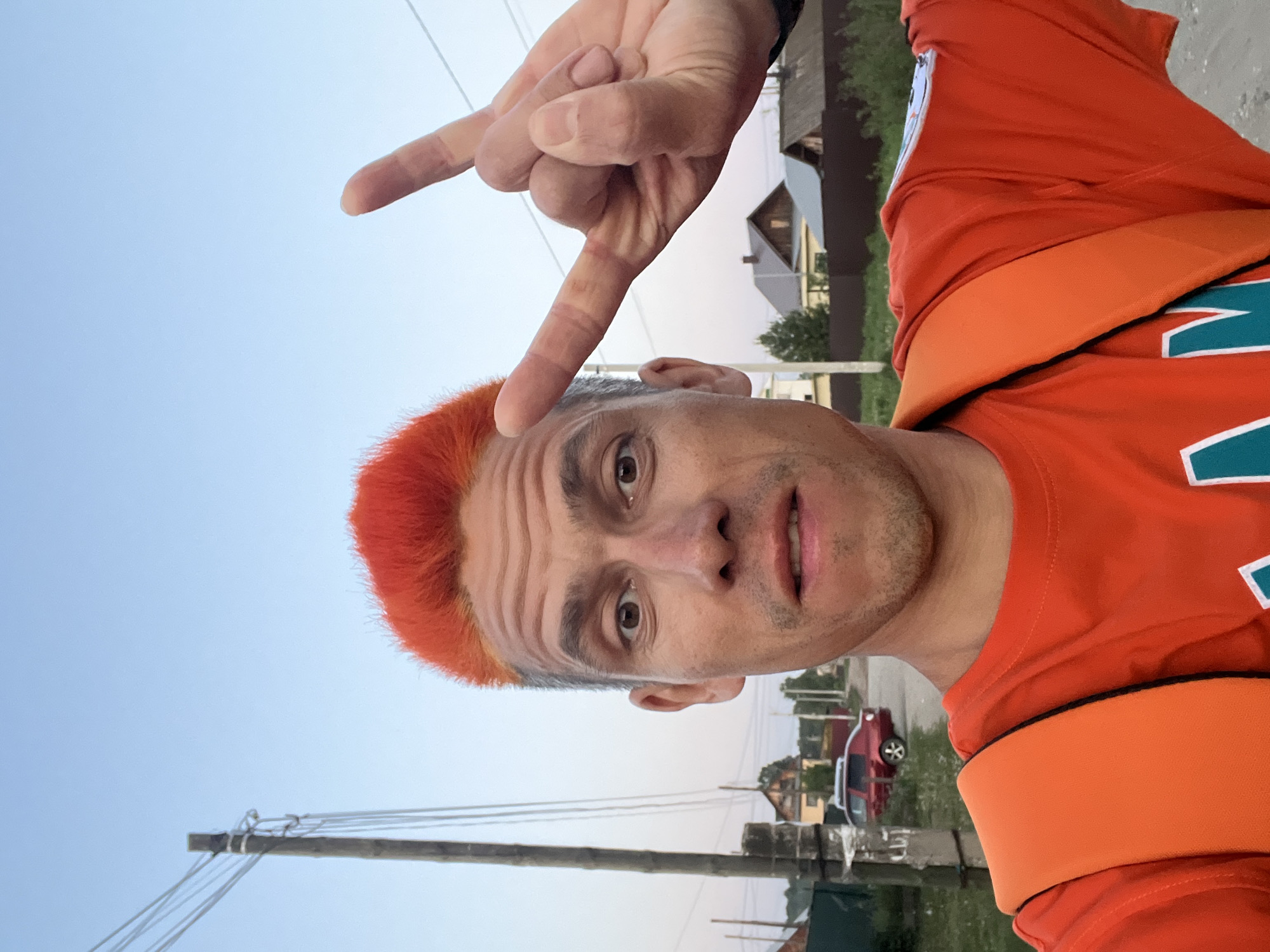 A man poses for a selfie in Chaikovsky, Russia. He is wearing an orange shirt with a Miami Dolphins logo. He has short, red hair and is making a "rock on" hand gesture above his head. In the background, there are houses, power lines, a red car, and some greenery. The sky is light blue.