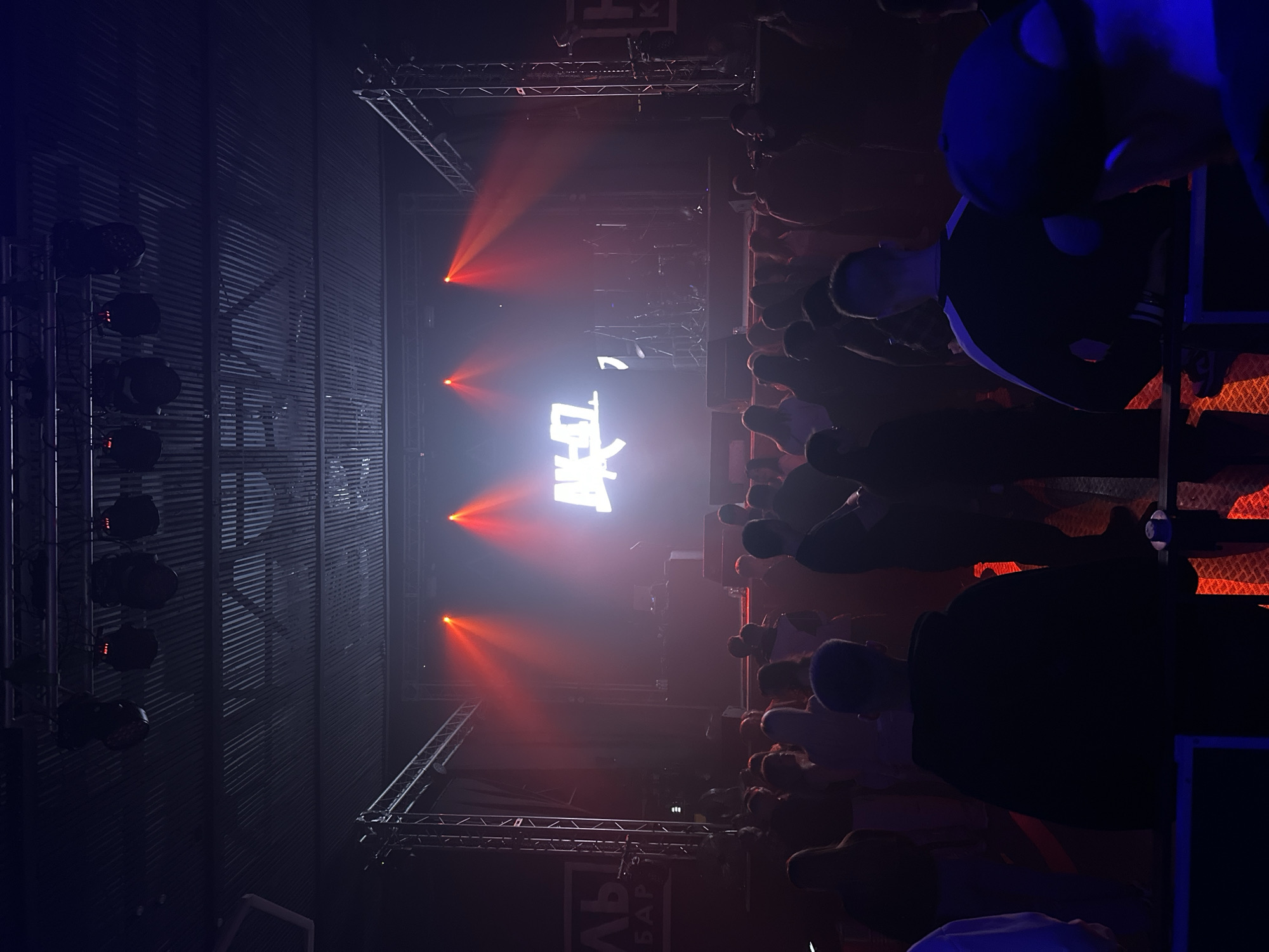 A vertical, eye-level shot shows a concert audience at a venue in Izhevsk, Russia. Red beams of light from the stage illuminate the crowd, directed toward a large, glowing white logo. To the left of the stage, a wall features vertical bars with mounted lights. The audience consists of numerous individuals, their heads and shoulders visible in the dimly lit space. Some audience members are holding up their cellphones. Parts of what appears to be a metal bar or scaffold are in the foreground and right side of the image. A sign with Cyrillic text is in the foreground.