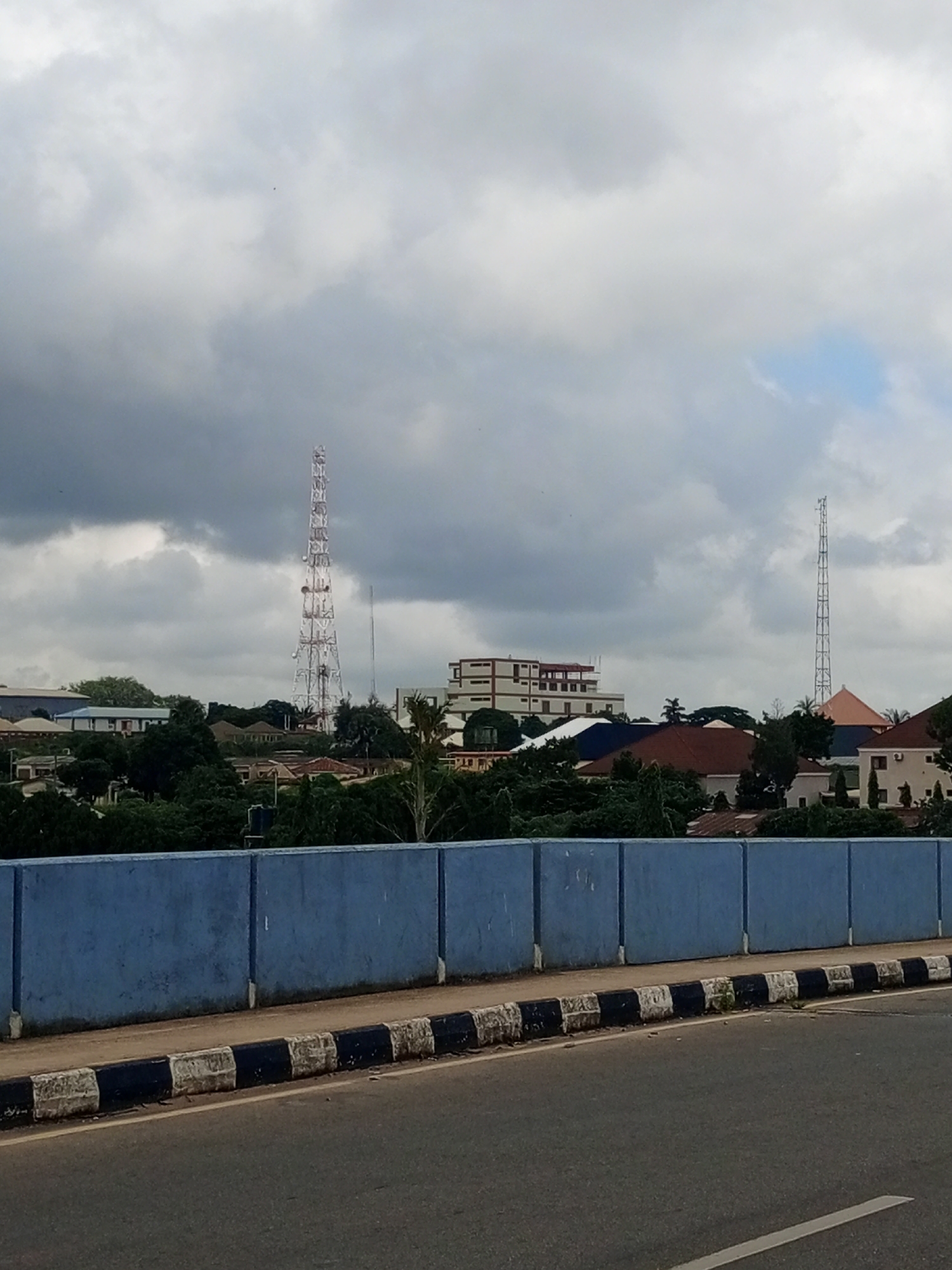 The image captures a cityscape of Jos, Nigeria, on a cloudy day, likely taken from a bridge or elevated roadway. In the foreground, a blue concrete barrier separates the roadway from the view beyond. The roadway itself is visible, with a black and white striped curb along its edge.
Dominating the middle ground is a dense cluster of trees, characteristic of urban greenery. Amongst the trees, various buildings are visible, including residential houses with red and brown roofs, and larger structures like a multi-story building with white and red sections. The skyline is punctuated by two tall communication towers, one very prominent and lattice-like on the left, and another thinner one further to the right. The sky is overcast with dark grey clouds, suggesting an impending or recent rain. There are no people or discernible actions depicted in the image. The scene is static and presents a typical urban view in Jos.