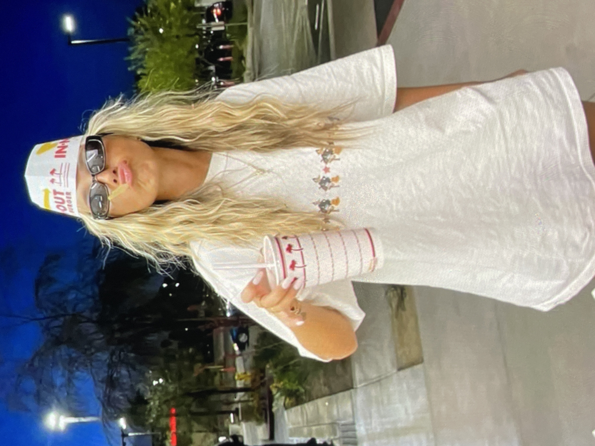The image shows a woman with long blonde hair wearing a white In-N-Out Burger paper hat and sunglasses. She is also wearing a white t-shirt with a small design on the chest. She is holding a red and white In-N-Out Burger drink cup with a straw in her right hand. The background appears to be a dimly lit outdoor area with trees and streetlights, suggestive of an evening setting. The location in Harmanli, Bulgaria is not discernible from the image.