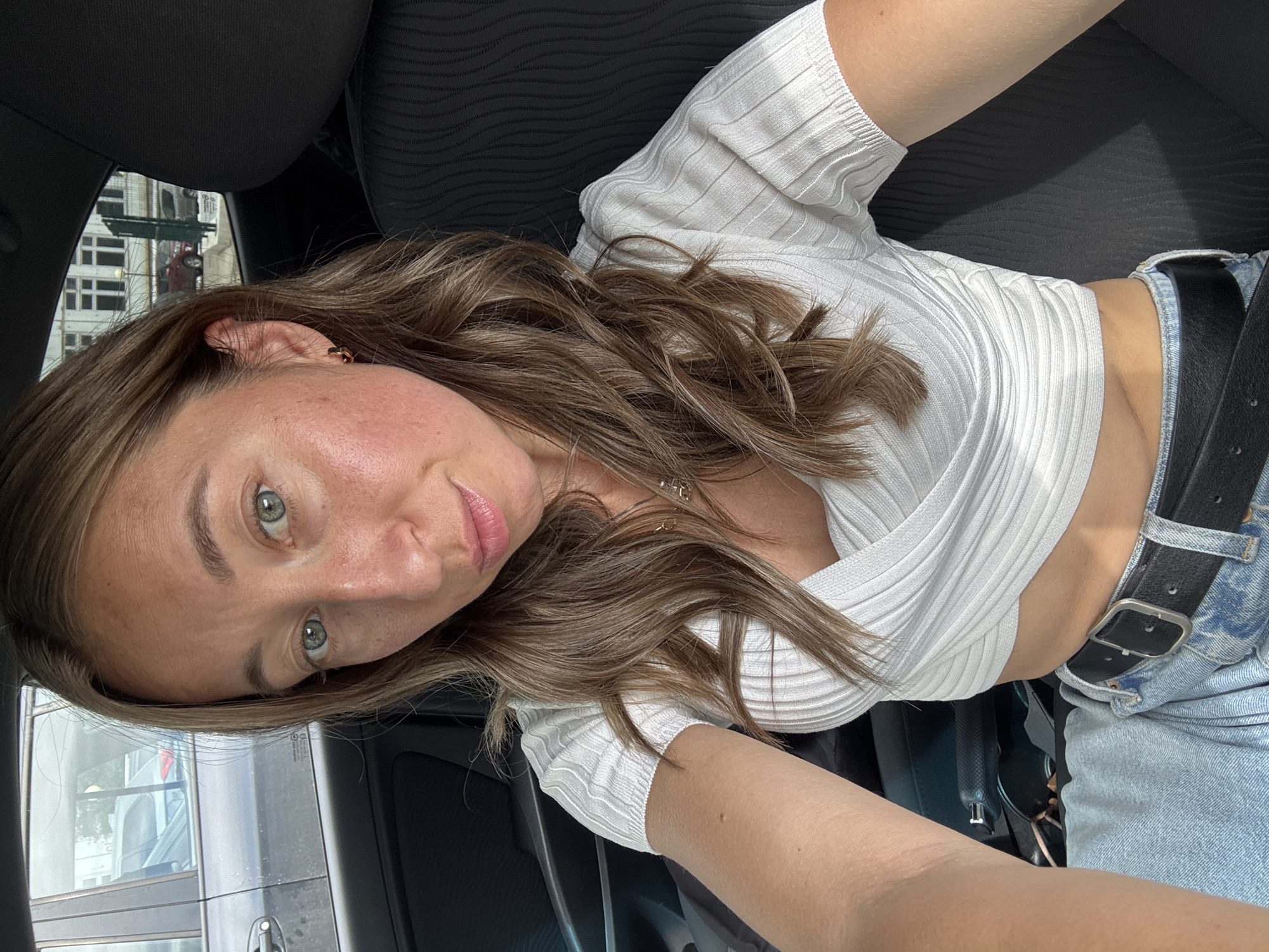 The image is a selfie, rotated 90 degrees clockwise, depicting a woman in the passenger seat of a car in Chaikovsky, Russia. The woman has medium-long, wavy brown hair, fair skin, and light-colored eyes, with a slightly pensive, calm expression as she looks directly at the camera. Her cheeks show some natural redness. She is wearing a light-colored (likely white) ribbed crop top with a wrap-around design, light-wash denim jeans, and a black belt. A small gold-colored hoop earring is visible on her right ear, and a thin gold-colored necklace is partially seen around her neck.
The car interior features dark-colored seats with a textured pattern, and a black seatbelt crosses her lower torso. Through the car window, parts of light-colored urban buildings with multiple windows are visible in the background, along with a hint of a red vehicle. The bright, clear lighting suggests it is daytime. No visible text is present in the image.