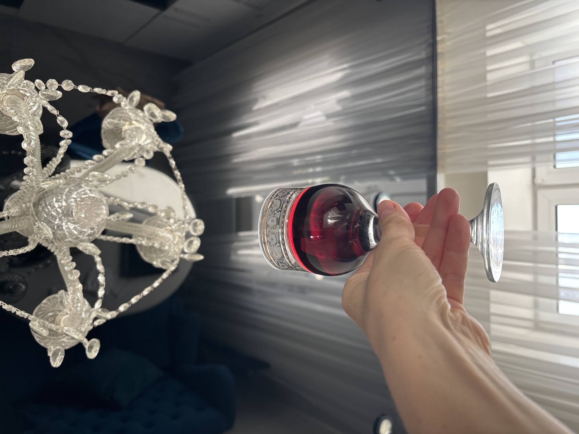 A hand holds a stemmed glass filled with dark red liquid, possibly wine. The glass has an ornate silver band with a floral pattern around its rim. In the background, a crystal chandelier with intricate details is visible, casting light and reflections. The scene appears to be indoors, with blinds covering a window in the far right, suggesting daytime. The overall ambiance is somewhat formal, perhaps a dining room or living area. The image is oriented vertically.