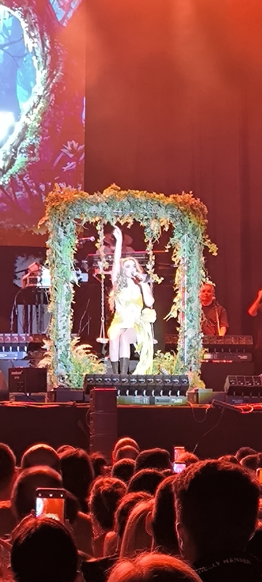 Here's a description of the image:
**Location:** The image appears to be taken at a live performance in Cerdanyola del Vallès, Spain.
**Subject:** A female performer is center stage on a swing that is suspended within a square frame decorated with greenery. She's wearing a yellow dress, dark boots, and is holding a microphone with one hand raised.
**People:** Several audience members are visible in the foreground with some holding up their phones. There are also other musicians and crew members behind the stage.
**Objects:** The scene includes musical instruments such as keyboards, speakers, and other stage equipment. The focal point is the swing and frame decoration.
**Action:** The main action is the female performer singing and gesturing. The audience is likely watching and recording the performance.
**Environment:** The scene is a concert setting with red lighting. The stage has multiple levels.