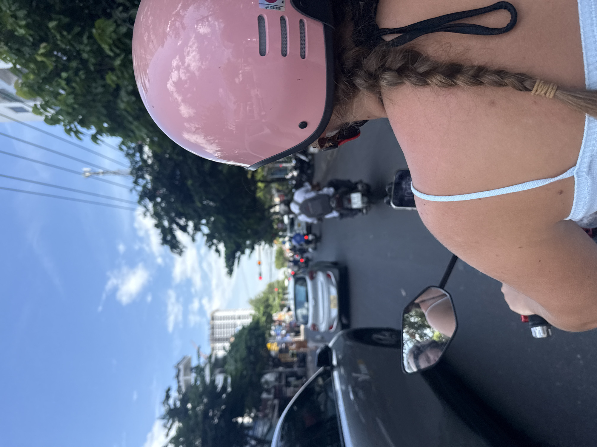 The image shows a person riding on the back of a motorcycle in the city of Nha Trang, Vietnam. The person is wearing a pink helmet and a light blue tank top, with their hair in a braid. The motorcycle is in traffic, with cars and other motorcycles on the road. The background shows buildings, trees, and a blue sky with clouds. The scene appears to be on a sunny day.