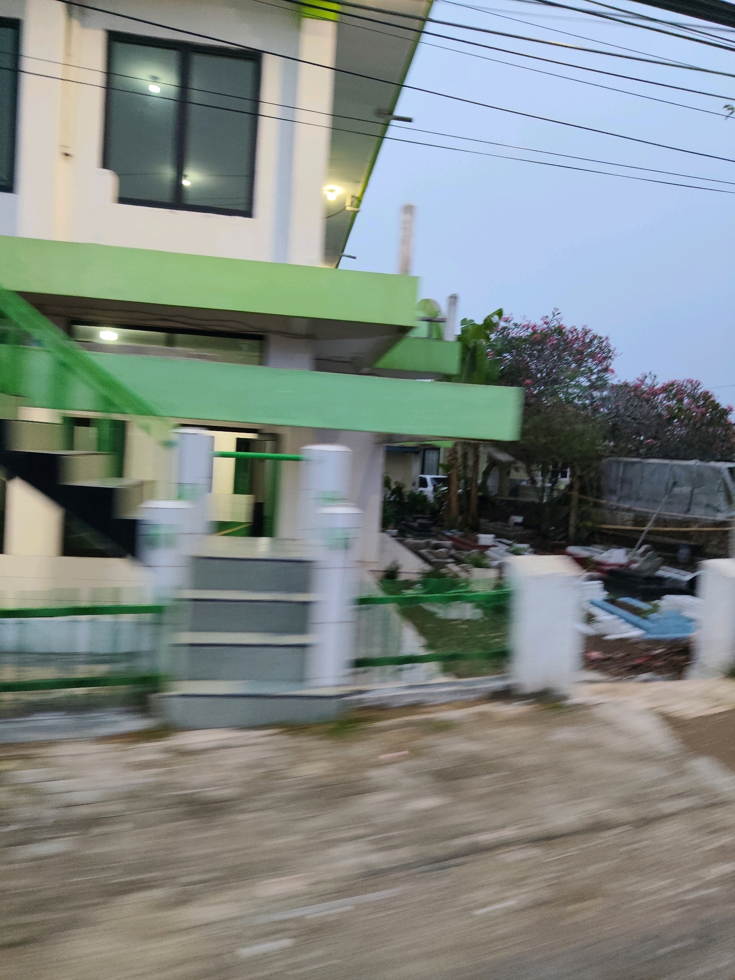 A blurry, motion-captured image shows the exterior of a white building with green trim, likely in Kaduagung Timur, Indonesia. The building has multiple levels and dark-tinted windows, some of which have lights on inside. A green railing runs along the lower part of the building, and a set of grey stairs with a green railing leads up to an entrance. Beyond the building, there is a yard with some vegetation and what appears to be debris or construction materials, including blue plastic sheeting. A white vehicle is partially visible in the background. Overhead, power lines are strung across the sky. The overall impression is of a residential or commercial property in an outdoor setting.