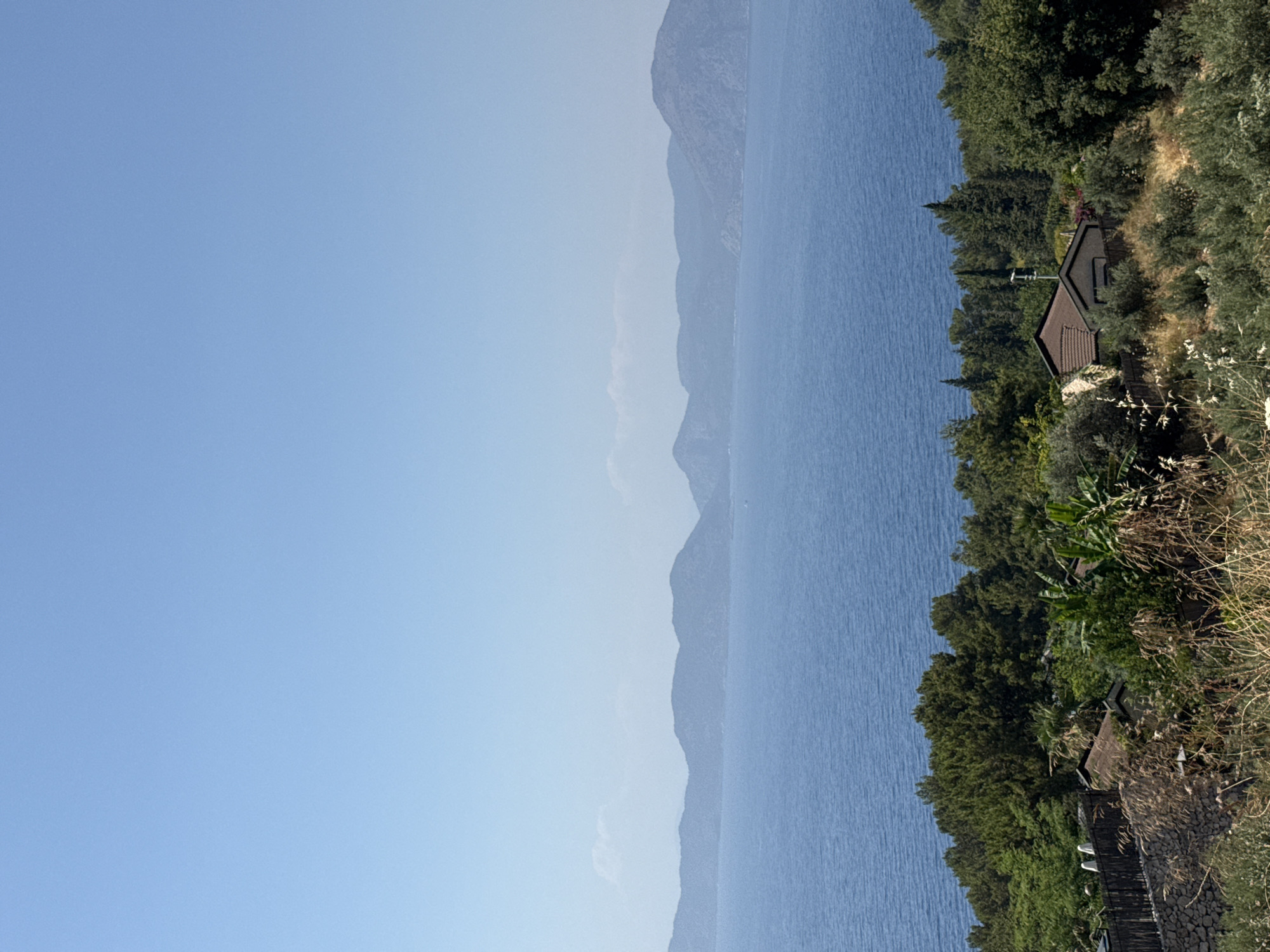 A panoramic view captures a coastal landscape under a clear, pale blue sky. In the foreground, a lush hillside is covered with dense green trees and bushes, with dry, golden grasses interspersed. A rustic stone wall and wooden fence are visible on the left, bordering a small cluster of residential structures. One building has a reddish-brown tiled roof and a dark garage door. In the middle ground, a vast expanse of deep blue water stretches out, calm and unbroken. Beyond the water, a series of hazy, dark blue mountains form the horizon, their undulating shapes softened by the distance. The overall impression is one of serene natural beauty and peaceful habitation, likely situated in a region like Fethiye, Turkey, known for its scenic coastlines and mountainous terrain.