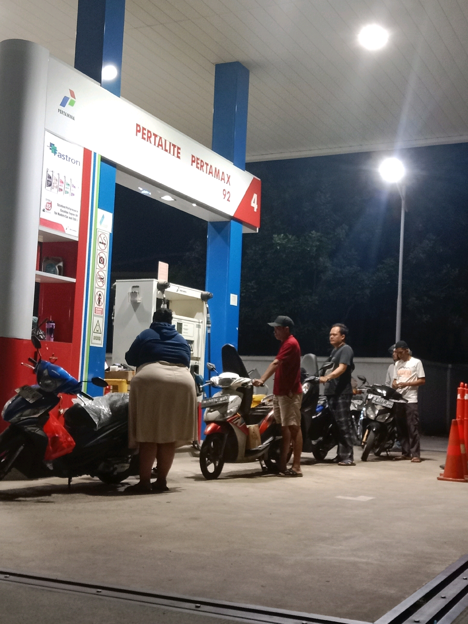This image depicts a scene at a Pertamina gas station in South Tangerang, Indonesia, during the nighttime. The gas station features blue and white signage, with "PERTALITE" and "PERTAMAX 92" prominently displayed. Several people are present, interacting with motorcycles. In the foreground, a woman in a dark blue hoodie and a beige skirt stands facing away from the camera, near a motorcycle covered with plastic sheeting. To her right, two men stand next to motorcycles. One man in a red shirt and khaki shorts is on the left, and another man in a dark shirt and plaid pants is to his right. Further to the right, a third man in a white shirt and dark pants is also near a motorcycle. The gas station canopy is illuminated by bright overhead lights, casting reflections on the concrete ground. The background shows dark foliage, indicating an outdoor setting at night. Several orange traffic cones are visible on the far right side of the image.