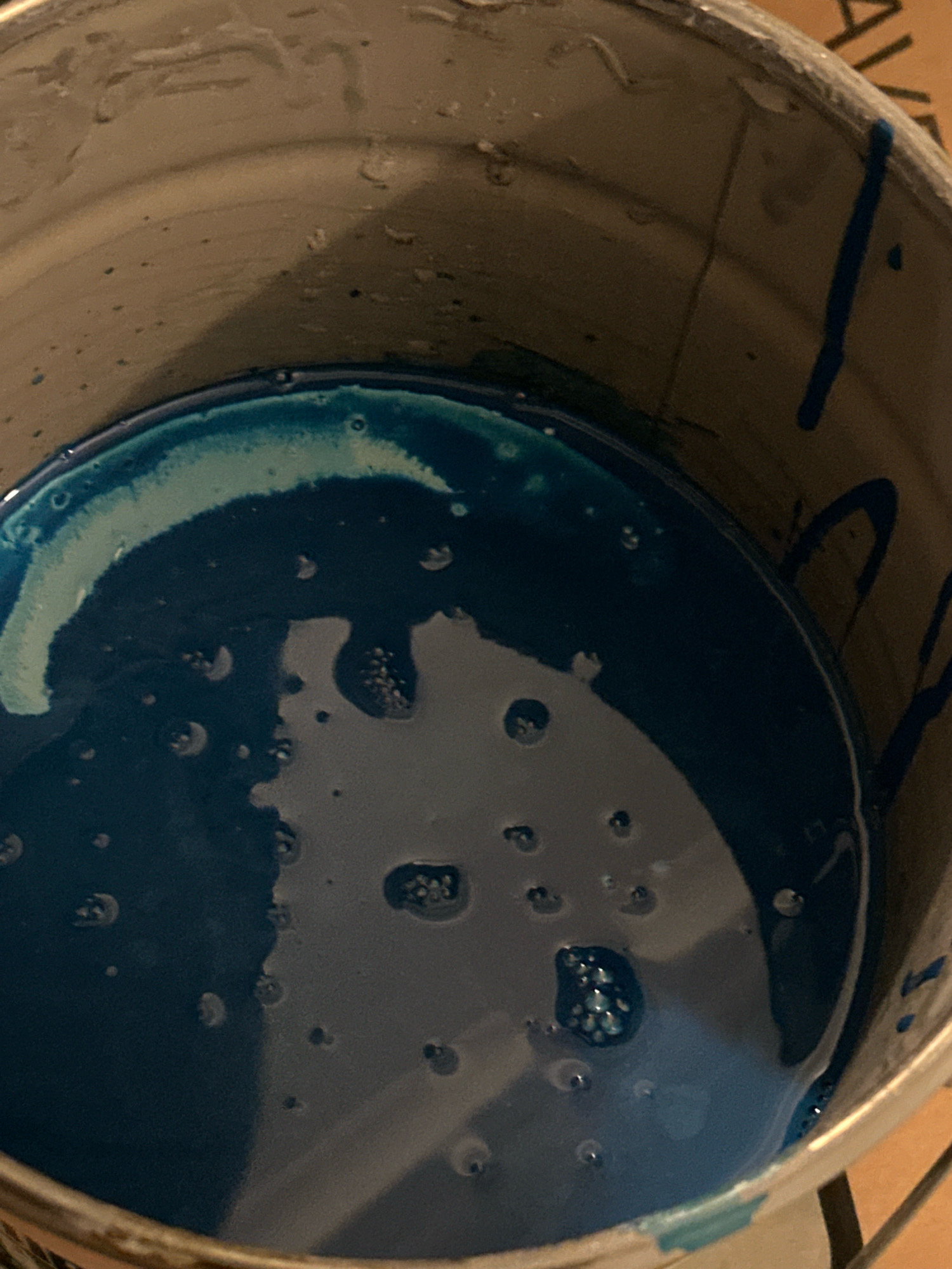 The image shows the interior of a metal can containing a dark blue liquid, possibly paint or resin. The liquid has a glossy surface with some visible bubbles and a lighter, foamy edge near the rim of the can. Blue streaks of the same liquid are visible on the inside walls of the can, suggesting it has been recently used or stirred. The can's interior is worn and shows signs of previous use. The background is out of focus but appears to be a light brown, possibly a wooden surface. There are no people or identifiable objects from Nevinnomyssk, Russia, in the image. The scene is a close-up view of the contents and interior of the container.