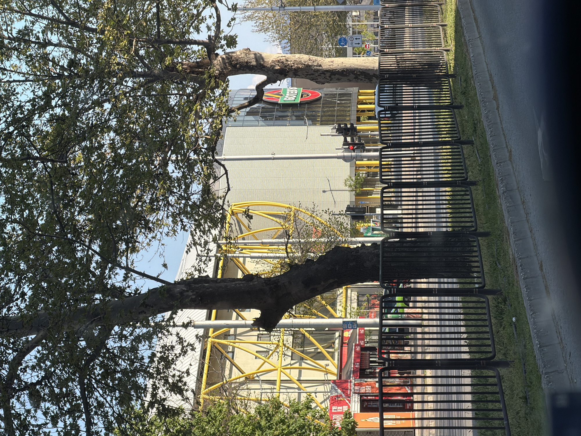 Here is a description of the image:
The image depicts a street scene in an urban area. In the background, there is a building with a distinctive sign that seems to display the word "GOTENBERG" in a red oval with a green banner. A yellow geometric structure, possibly part of a building or construction, is visible. To the right of the frame, there are several black metal barriers lining the sidewalk. The scene is partially obstructed by a large tree with lush foliage, which dominates the left side of the image. The photograph is taken from a low angle, perhaps from a moving vehicle. A street and signs are barely visible to the right. The exact location within the city of Unknown, Unknown, is difficult to determine definitively, but the signage on the building suggests a possible cultural or commercial identity related to the name shown.