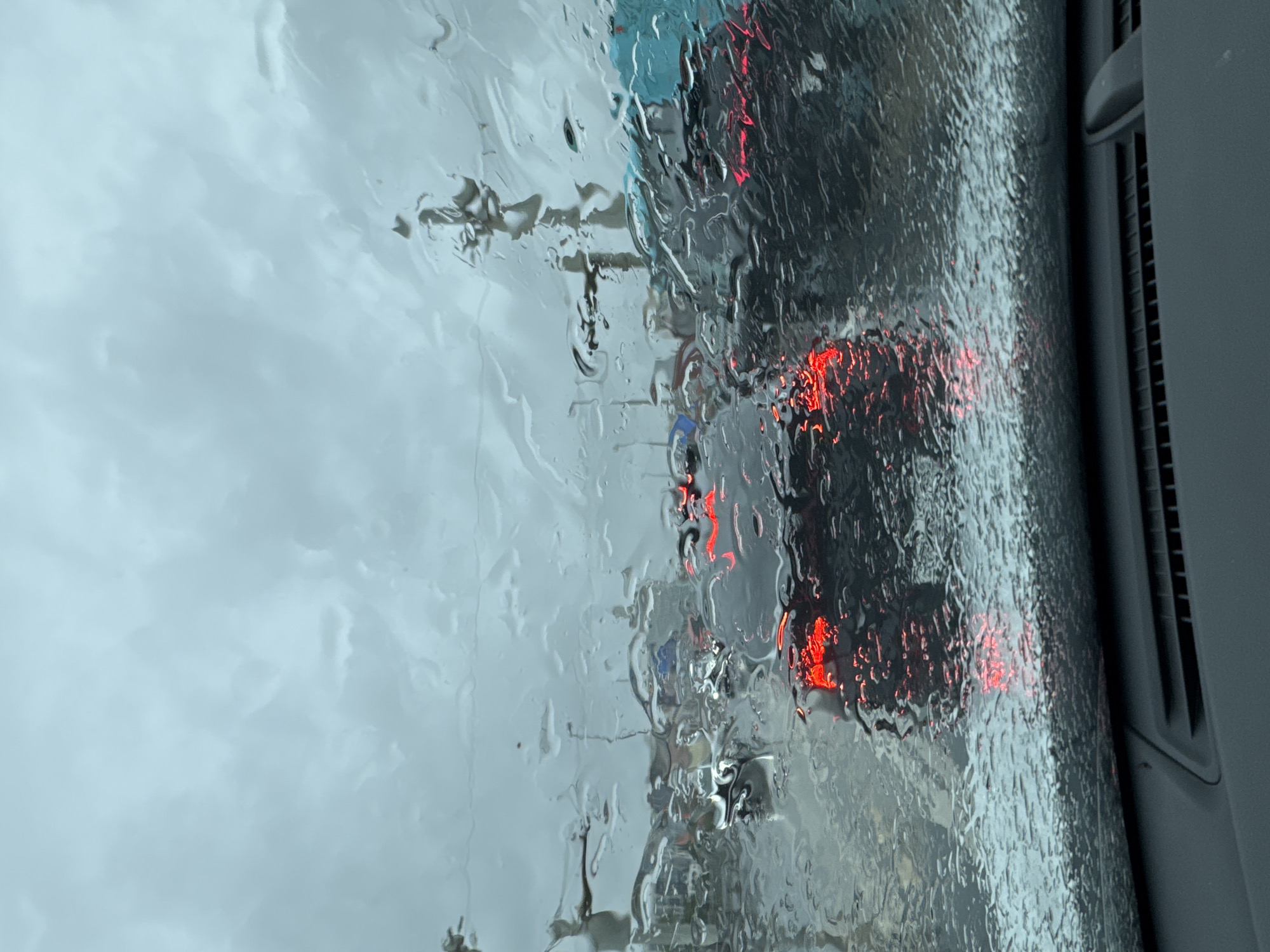 The image shows a view from inside a vehicle looking through a rain-streaked windshield at traffic. The sky is overcast and grey. Red taillights from vehicles in front are visible through the rain. Buildings and streetlights can be seen in the distance. The foreground shows the dashboard of the vehicle. The scene appears to be a traffic jam on a wet, cloudy day.