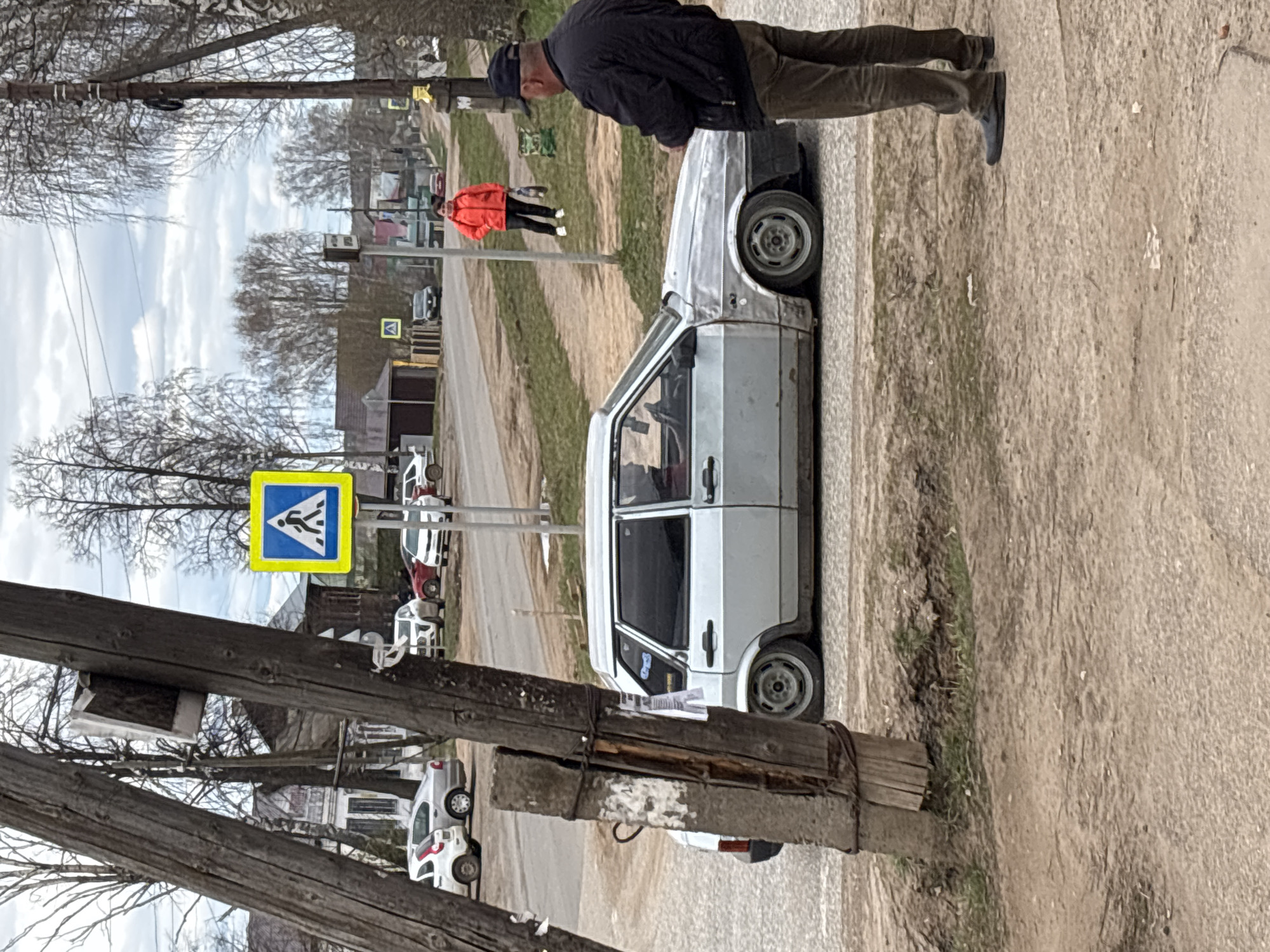 This image, rotated 90 degrees clockwise, depicts a rural or semi-rural street scene in what appears to be Russia. The primary subject is a man wearing a dark jacket and a cap, standing beside a white, two-door sedan, likely a Lada Samara or similar model, which is parked on a dirt and gravel shoulder. The car shows signs of wear and appears to have a lowered suspension.
In the midground, further down the unpaved road, another individual wearing a bright orange jacket is visible. The road itself is made of dirt and gravel, with grass and sparse vegetation on its sides. Several other cars are parked or moving in the background. Prominently displayed near the center-left is a yellow square road sign with a blue triangle inside, featuring a white pedestrian figure, indicating a pedestrian crossing.
The sky is overcast, and the trees scattered throughout the scene are bare, suggesting either early spring or late autumn. In the left foreground, two rustic wooden utility poles are visible, one angled across the bottom left of the frame. The overall setting is unpaved and sparsely developed, consistent with a village or small town environment. There is no legible text visible other than the symbol on the road sign.