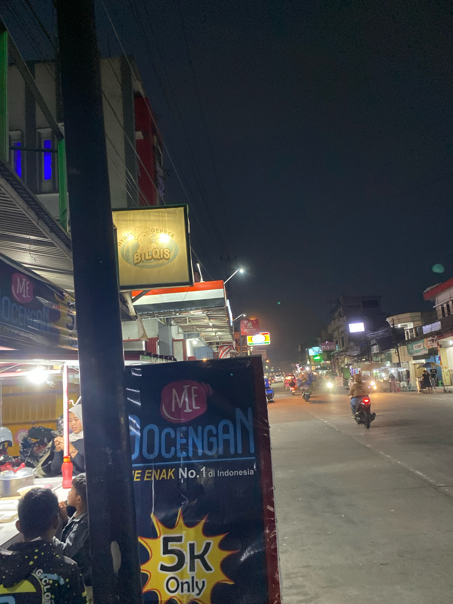 This is a nighttime photograph of a street scene in Kertek, Indonesia. A dark pole is in the foreground, obscuring part of the view. To the left, a food stall is visible with a woman in a headscarf preparing food. Several people are gathered at the stall. A large banner in front of the stall advertises "JOCENGAN" and "5K Only". Above the stall, a sign reads "AYAM GEPREK BILQIS". The street is lined with buildings and other stalls, with several motorcycles and a few cars visible on the road. The overall impression is of a busy street food area at night.