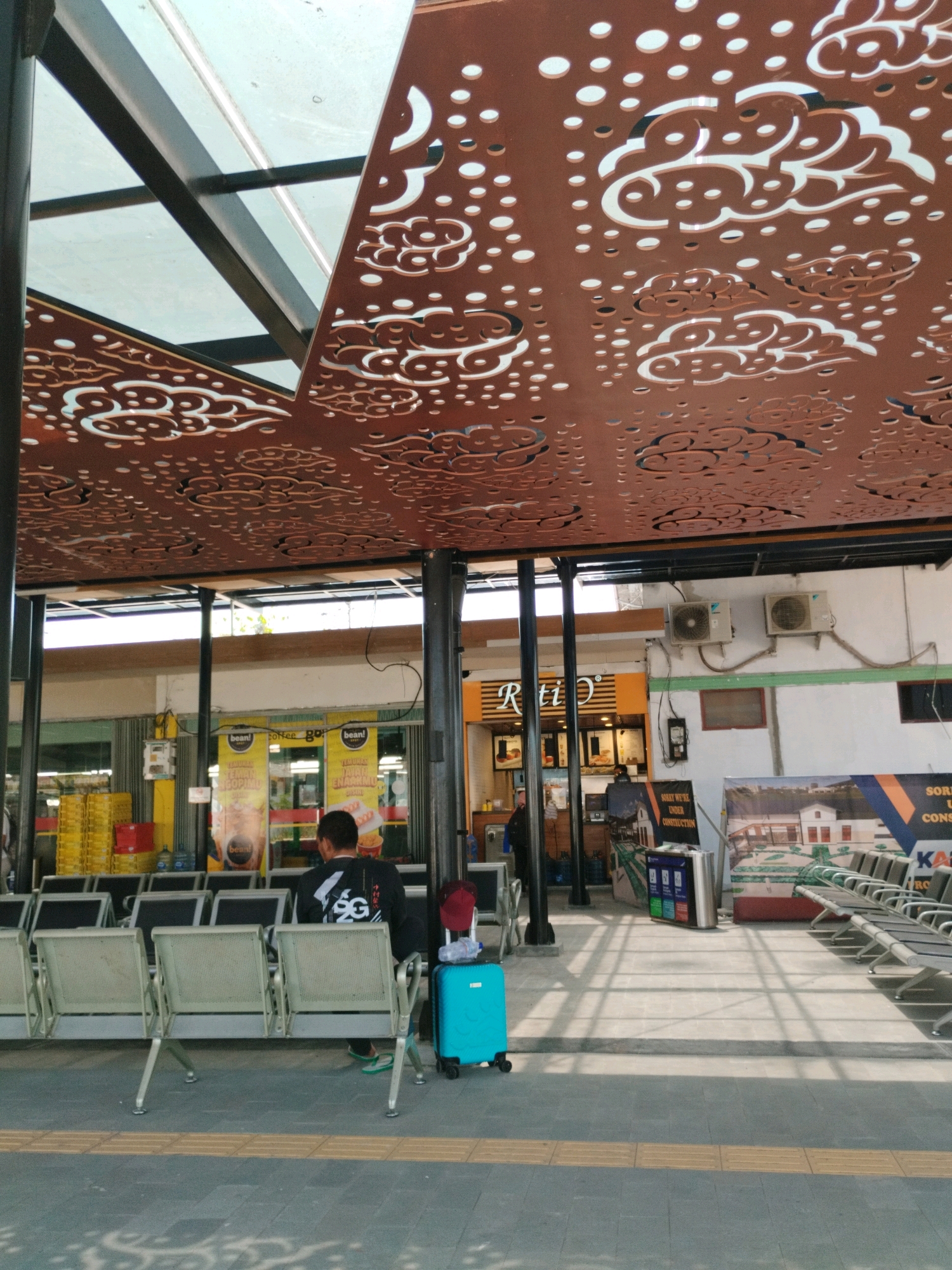 The image captures a covered waiting area, likely at a public transport station in Jember, Indonesia. A person, viewed from behind, is seated on a light-colored bench, facing away from the camera. They are wearing a dark-colored long-sleeved shirt with a white graphic and dark pants. Next to them is a bright blue suitcase and a red cap resting on a nearby seat. Rows of identical benches are arranged in parallel, stretching into the background. Overhead, a decorative ceiling made of perforated metal with a cloud motif casts dappled shadows on the floor. In the background, a small shop or cafe with orange and yellow signage is visible, featuring various advertisements. Two air conditioning units are mounted on the wall behind the shop. The overall environment appears to be a bustling, yet orderly, public space, common in urban areas like Jember.
