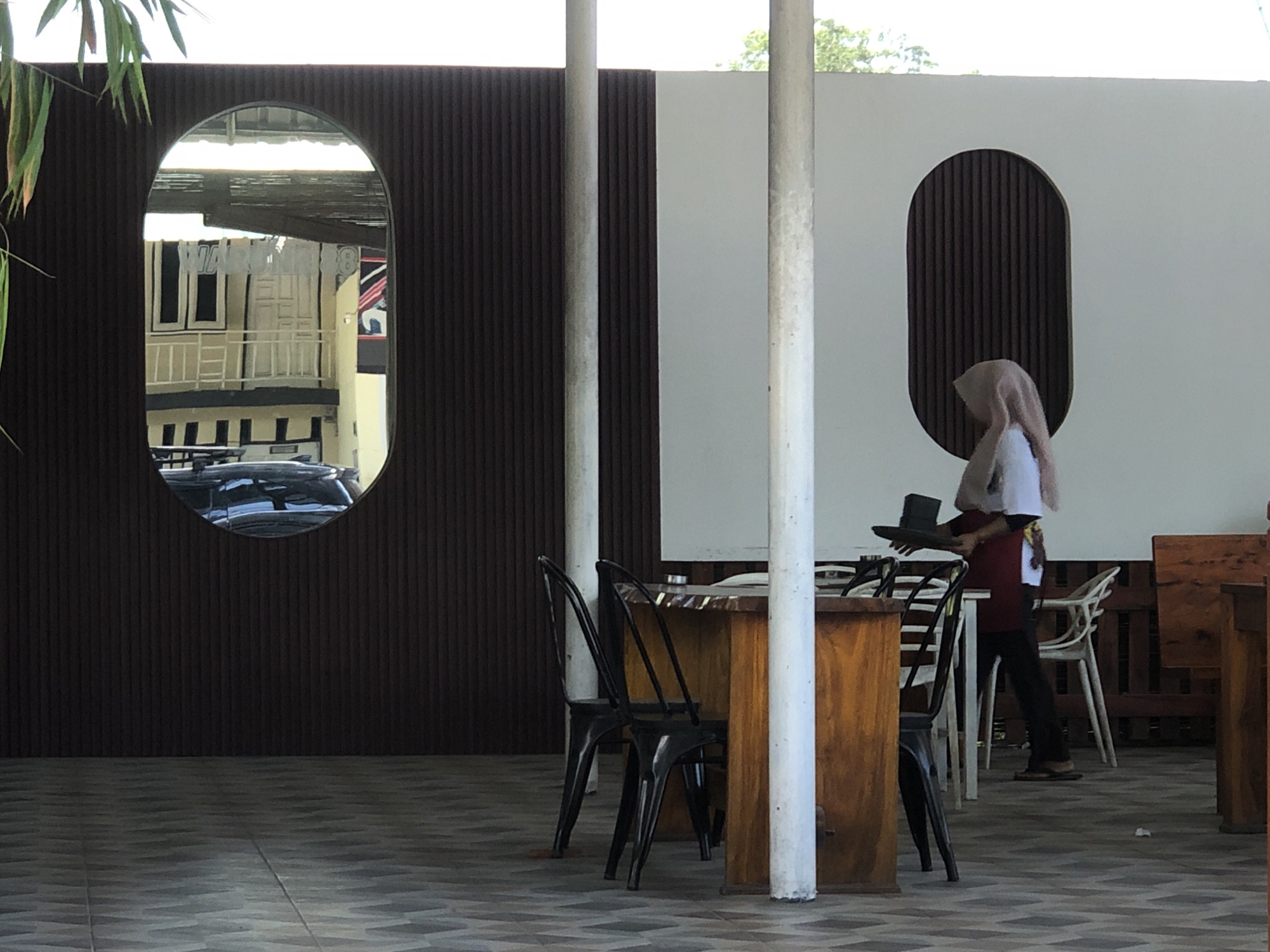The image depicts an outdoor seating area, likely a cafe or restaurant, with a reflective oval mirror mounted on a dark, ribbed wall. The mirror reflects buildings with balconies and a car. A person wearing a hijab and a red apron is walking away from the viewer, holding a dark hat and a tray. The scene is furnished with wooden tables and chairs, including black metal chairs and a white plastic chair. The ground is paved with patterned tiles. In the background, a white wall with a dark ribbed oval accent is visible. The location is identified as Teupinbatee, Indonesia.