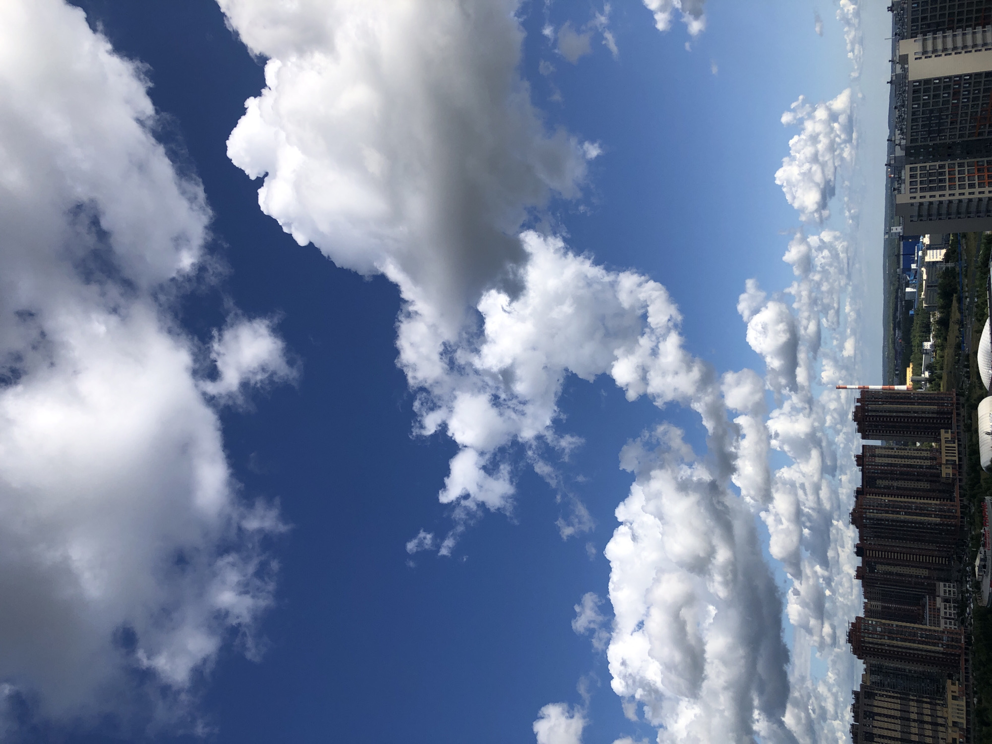 A high-angle, full shot captures a bright, sunny day in a city with a striking blue sky filled with voluminous white and gray clouds. In the foreground, a dense cluster of modern, multi-story apartment buildings with varied facades dominate the lower portion of the frame. These buildings are characterized by their uniform height, numerous windows, and balconies, suggesting a residential area. To the right of these buildings, a tall, red and white striped smokestack stands prominently, hinting at industrial activity in the distance. Beyond the apartment complexes, the cityscape recedes into a hazy horizon, with more scattered buildings and greenery visible. The overall impression is one of urban development under a dynamic sky.