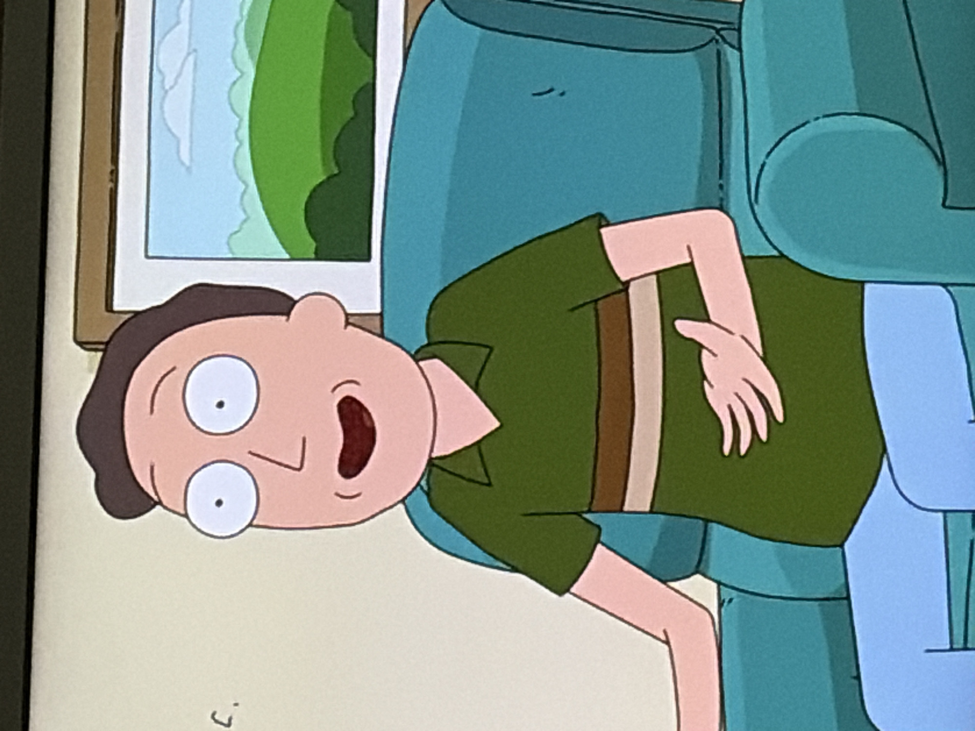 The image shows a medium close-up of a cartoon character, identified as Jerry Smith from the animated series "Rick and Morty". He is depicted sitting in a teal armchair, facing slightly to his left. Jerry has light skin, a round face with wide, surprised eyes, and a small mouth. He is wearing a green short-sleeved polo shirt with horizontal brown and white stripes across the chest, and light blue pants. His left arm rests on his lap, while his right hand is placed on his stomach. In the background, a framed landscape painting hangs on a beige wall. The painting depicts rolling green hills under a light blue sky with white clouds. A small, handwritten "C." is visible on the wall to the left of the frame. The scene appears to be indoors, within a residential setting.