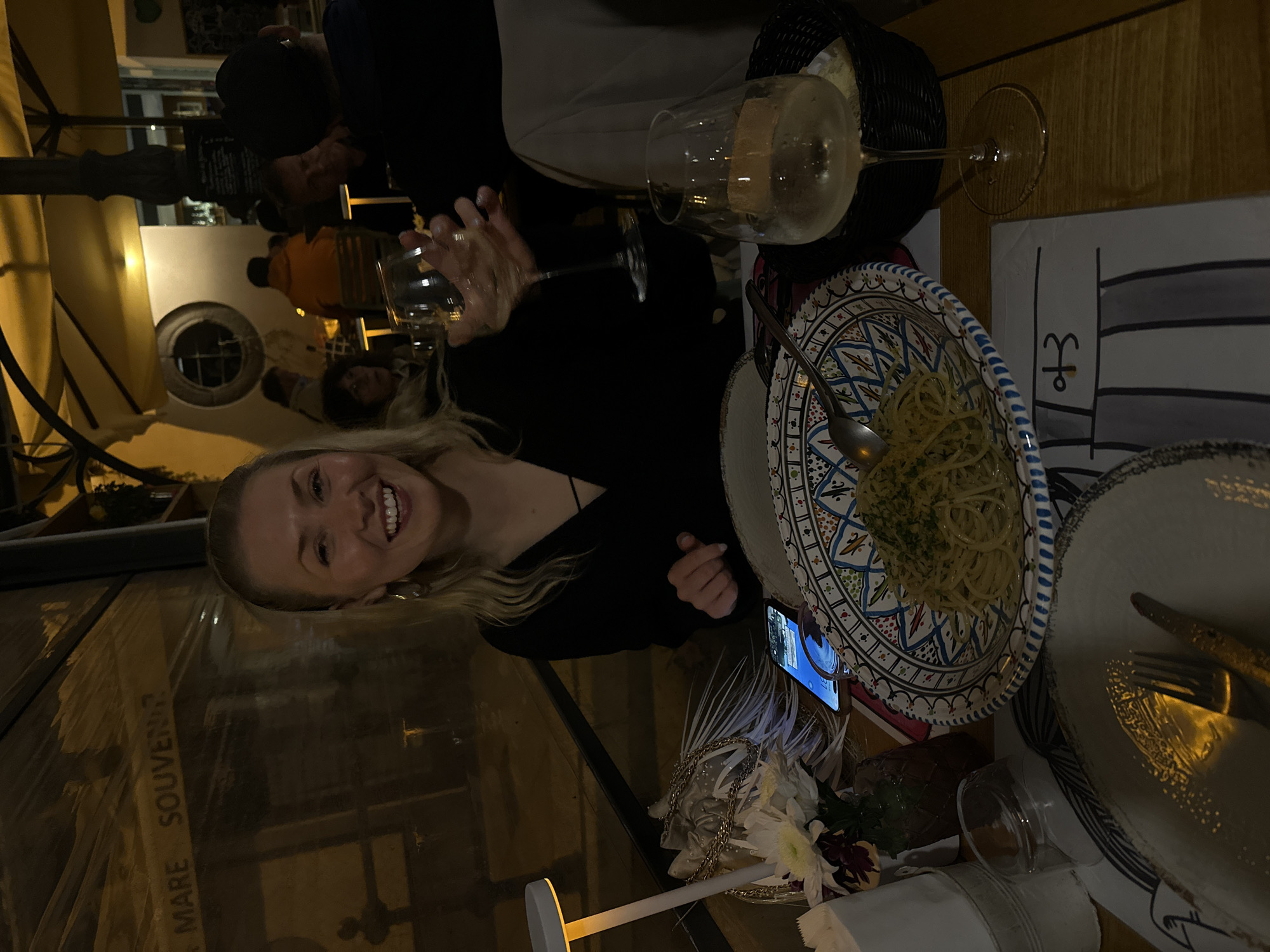 A woman with blonde hair is seated at a table, smiling at the camera, in what appears to be an outdoor dining area. She is wearing a black top. The table is set with a decorative plate of spaghetti, a wine glass filled with white wine, and a basket. A mobile phone is placed near the plate. A partially visible man with dark hair is behind her, seated at the same table. The dining area is under a yellow awning, and a window is visible in the background. The glass separating the dining area from the outside is etched with the words "MARE SOUVENIR." Based on the image content and the request for a description in Lipari, Italy, it can be inferred that this is likely a restaurant or cafe in the city.
