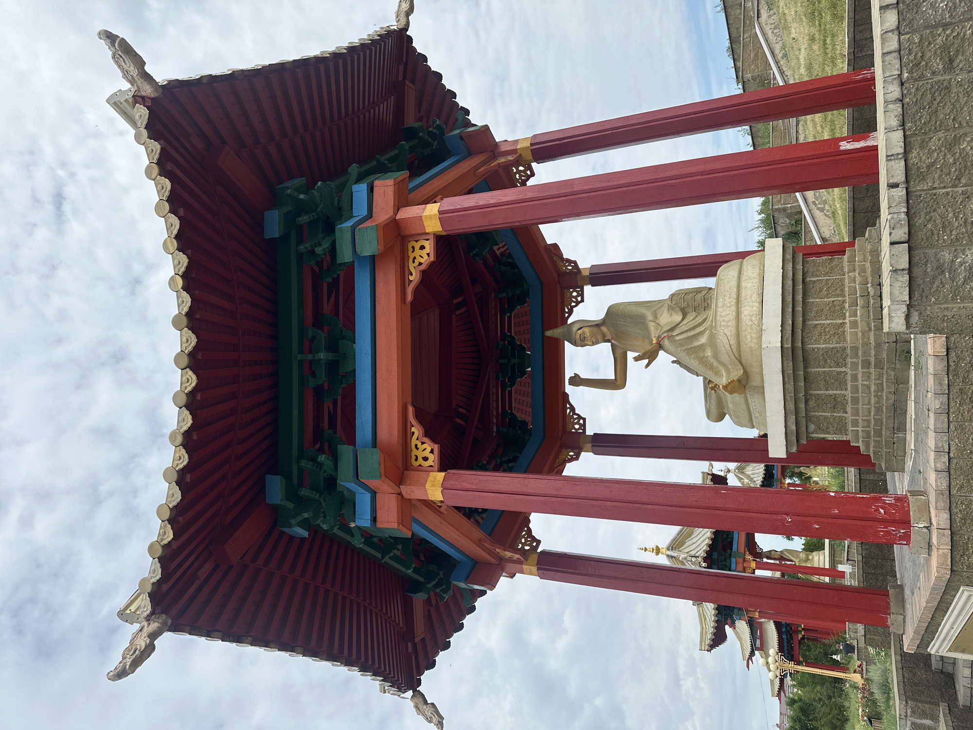 The image shows a Buddhist temple in Elista, Russia. The temple is octagonal in shape with a red and blue roof and red pillars. In the center of the temple, there is a gold statue of a seated Buddha. The statue is surrounded by a stone platform. In the background, there are other Buddhist temples and trees. The sky is cloudy. The image is taken from a low angle, looking up at the temple.