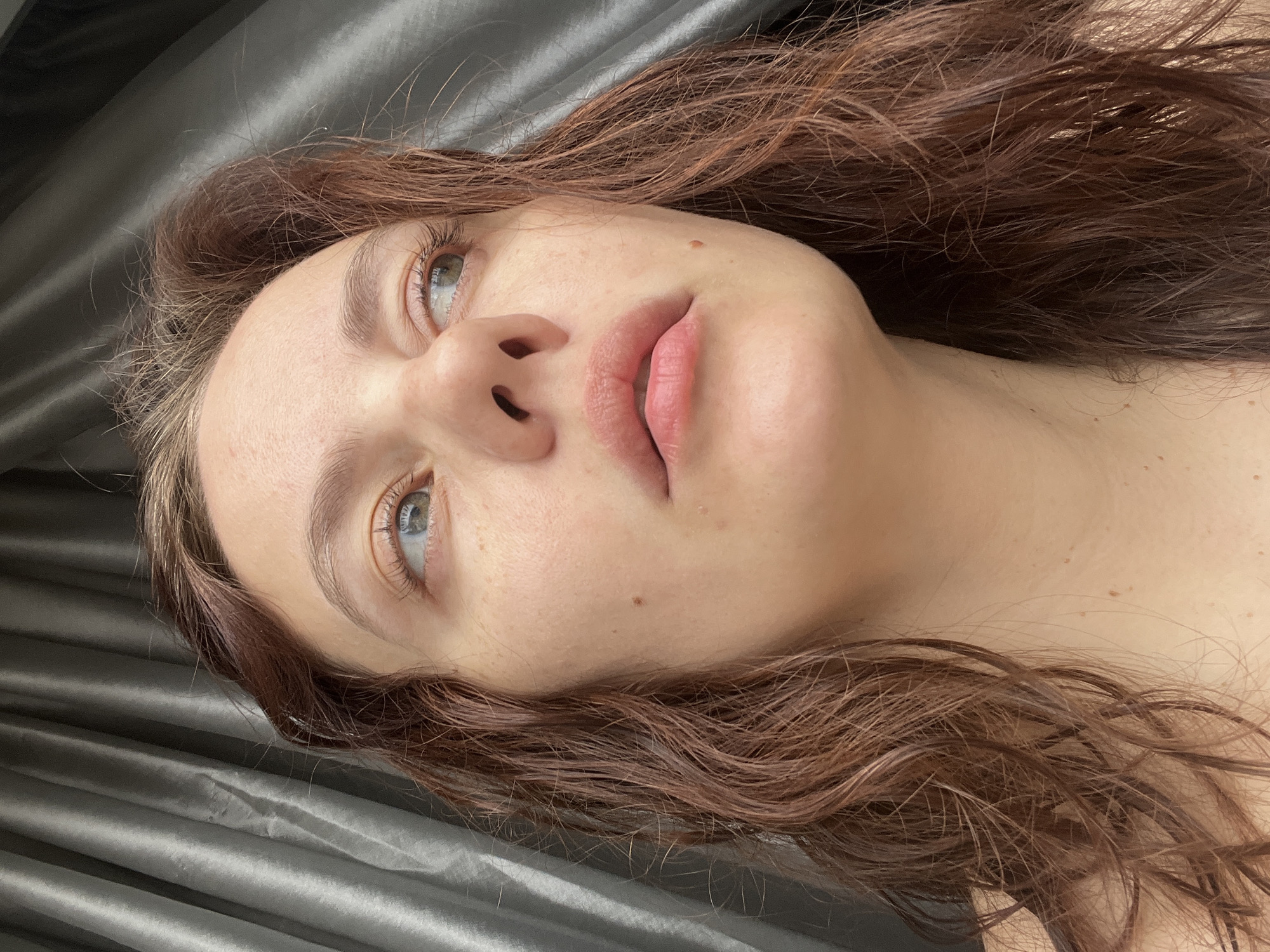 A close-up, slightly tilted, full-face portrait shows a young woman with fair skin and wavy brown hair lying on a gray, satin-like material. The woman's head is angled, with her face directed upwards and slightly to the left. Her eyes are open and appear to be light blue or green. She has natural, full lips and a visible mole on her cheek. The gray material beneath her appears to be a draped fabric, possibly a bedsheet. There is no indication of the location being Chaikovsky, Russia within the image itself.