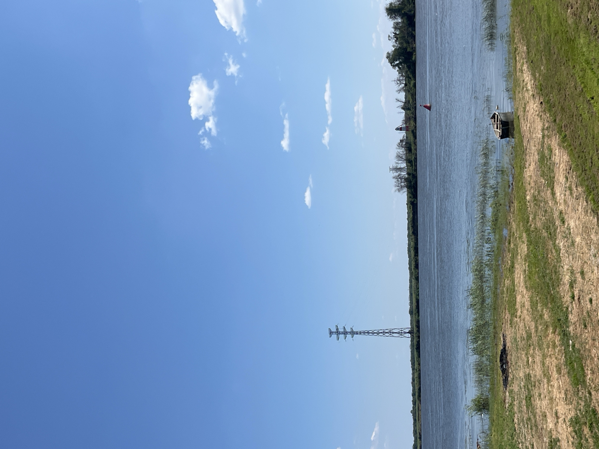 The image shows a wide river under a clear blue sky with scattered white clouds. On the far bank, a line of trees is visible, with a tall power line tower standing prominently to the left of center. Further to the right, some structures, possibly buildings, can be seen amongst the trees, along with a red and white buoy in the river. In the foreground, a sandy bank slopes down to the water, covered with sparse green grass and some patches of dry, yellowed grass. A small, dark rowboat is moored in the shallow water near the bank, and a dark, burnt area is visible on the sandy bank. The location is identified as the city of Krasnyye Barrikady, Russia.