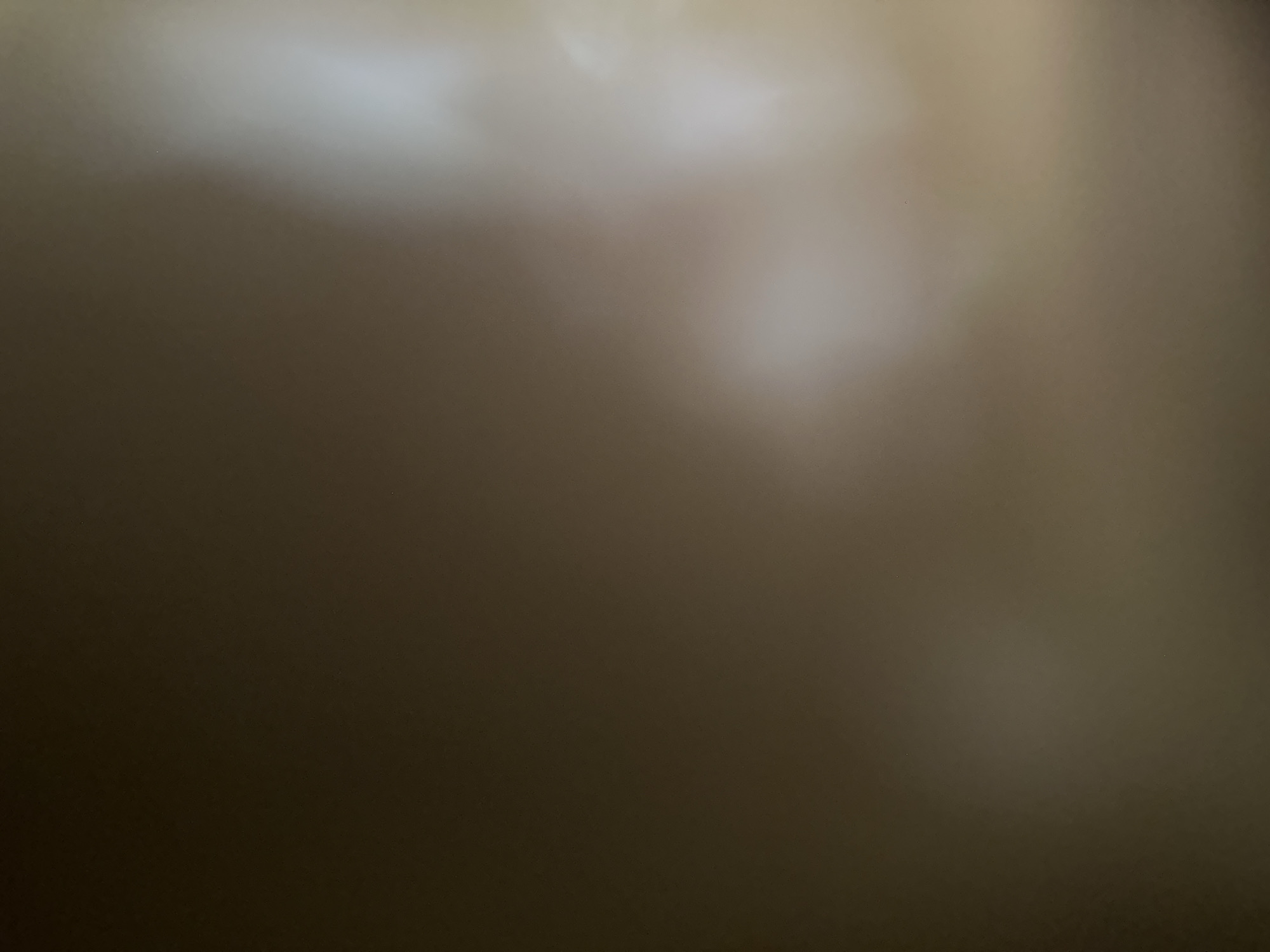 This is a close-up, out-of-focus image with a soft, diffused light source. The dominant colors are muted browns, grays, and off-whites, suggesting an indoor or dimly lit environment. There are no discernible subjects, objects, or a clear scene depicted. The image appears to be abstract due to its lack of focus and detail. It is not possible to determine the time of day, weather, location cues, emotions, or any visible text from this image.