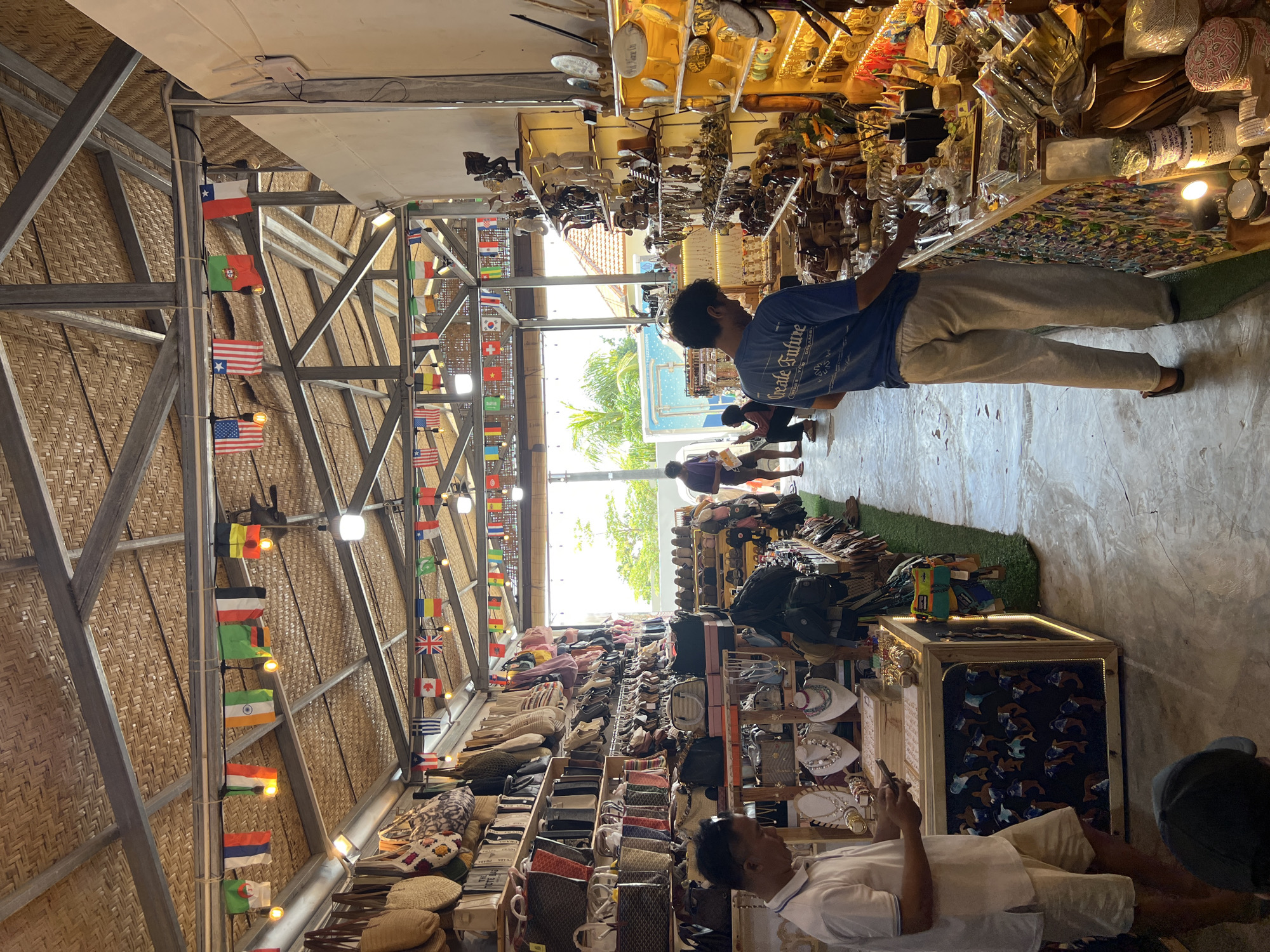 A long, vertical shot showcases an indoor marketplace in Kuta Selatan, Indonesia, under a bamboo ceiling adorned with a string of international flags.
On the right, a man in a blue t-shirt and light pants browses a merchandise display. Behind him, a few figures walk toward the bright outdoor entrance.
On the left, an individual in a white shirt is seated and focused on a phone. The space is filled with shelves displaying various merchandise, including bags, hats, and decorative items. The lighting is a mix of natural light from the entrance and artificial light illuminating the displays.