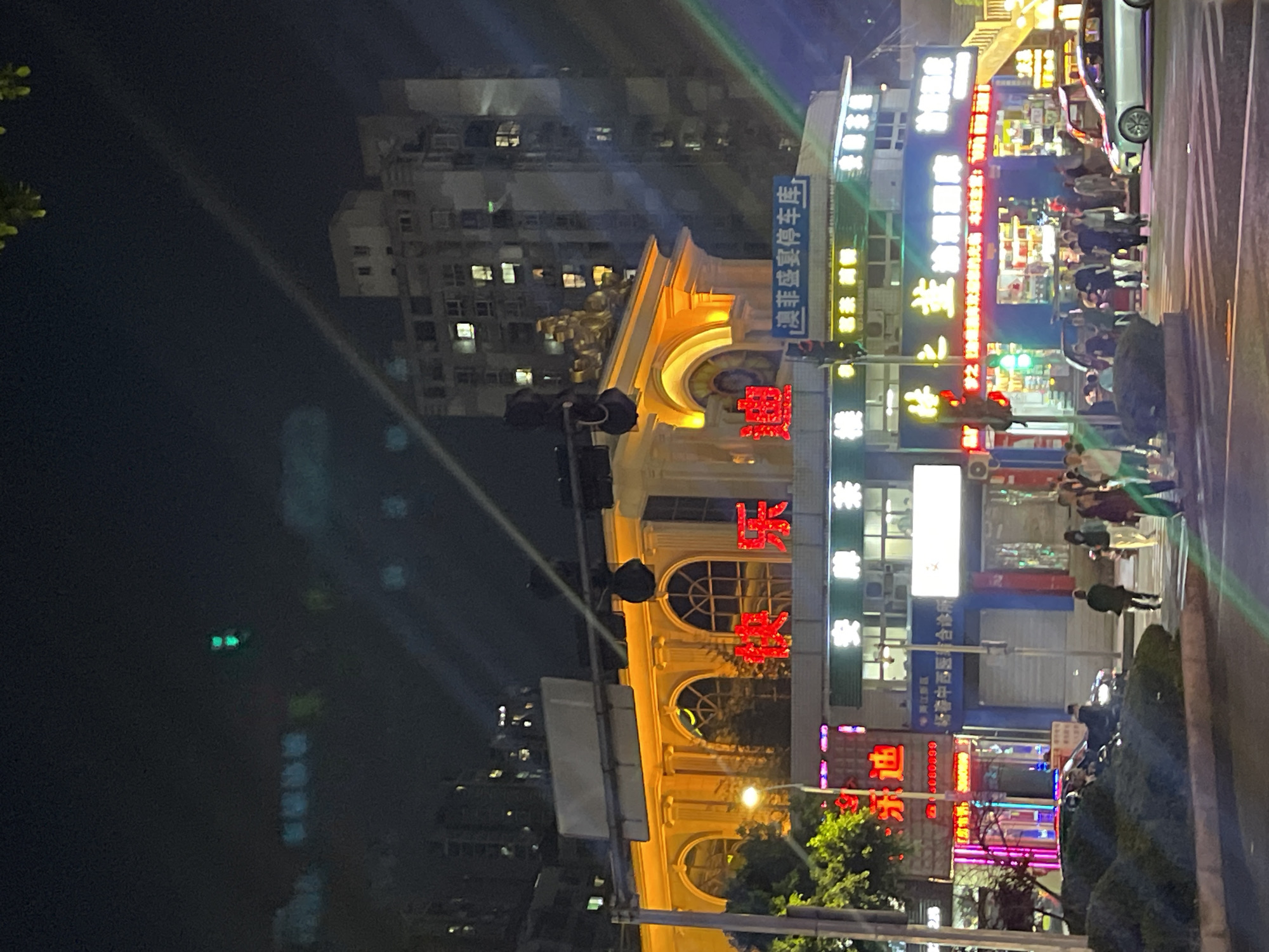This media file captures a vibrant night scene in an urban area, likely in Liangjiang, China. The setting is a bustling street lined with brightly lit commercial buildings and active pedestrian traffic.
In the mid-ground, a prominent building with a classical, ornate facade is illuminated in warm yellow light. Large red neon characters on this building clearly spell "澳菲盛宴" (Aussie Feast Banquet), indicating a restaurant or event hall. Above this, a blue sign displays "杨春中西医非合诊所" (Yangchun Chinese and Western Medicine Non-Integrated Clinic) and a phone number "03080899". Another sign mentions "澳菲盛宴停车库" (Aussie Feast Banquet Parking Garage).
To the right of this ornate building, a series of commercial storefronts feature various illuminated signs in red, blue, white, and yellow, creating a colorful glow. Below these signs, people are visible on the sidewalks and a crosswalk, suggesting active evening hours. A silver car is parked near the curb, and a scooter or motorcycle is also present amidst some greenery. Traffic lights displaying green indicate active road movement.
In the background, taller residential or commercial buildings with numerous illuminated windows rise against the dark night sky. The visible text also includes "黄江新区" (Huangjiang New District), providing a more specific location cue within the broader Liangjiang context. The overall atmosphere is lively and energetic, characteristic of a modern Chinese city at night.