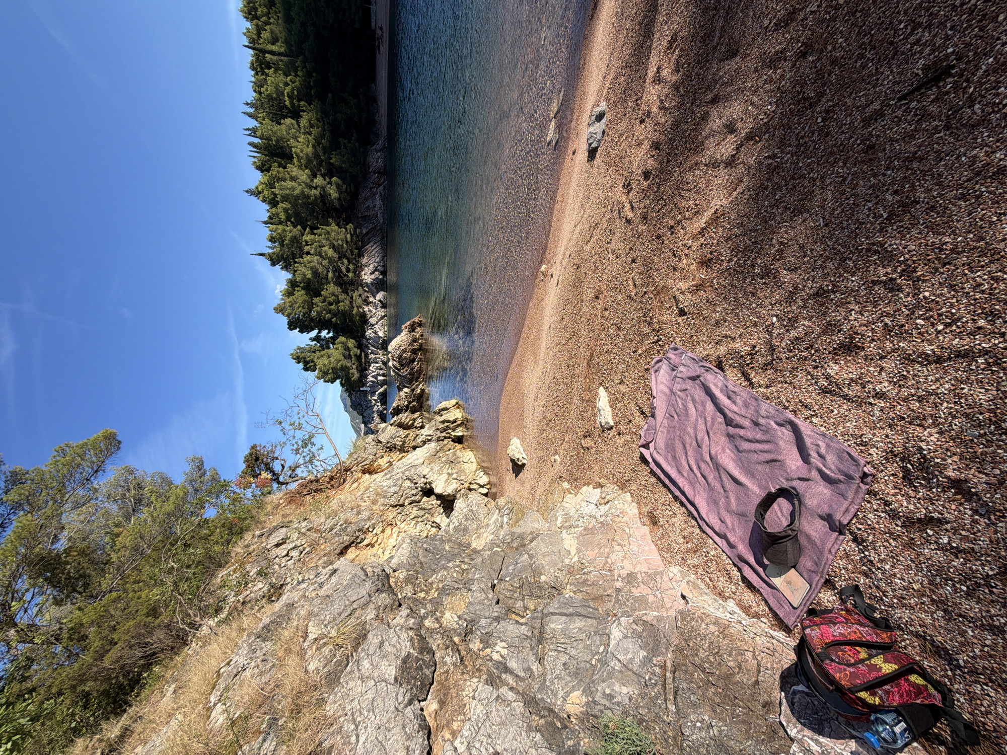 This image depicts a tranquil beach scene in Pržno, Montenegro, under a clear blue sky. The foreground is dominated by a rocky outcrop on the left, covered with dry grass and sparse vegetation, transitioning into a pebble beach. A purple towel is laid out on the beach, with a black mask and a phone placed on top of it. To the left of the towel is a colorful backpack, and a water bottle is visible within it. The beach slopes gently towards the water, which is a clear turquoise. The water is calm, with visible pebbles on the seabed near the shore. In the middle ground, several rocks are scattered in the water. The background features a dense line of green trees, primarily pine and cypress, lining the coastline, with a few more rocky inlets further in the distance. The overall atmosphere is peaceful and idyllic, suggestive of a secluded cove.